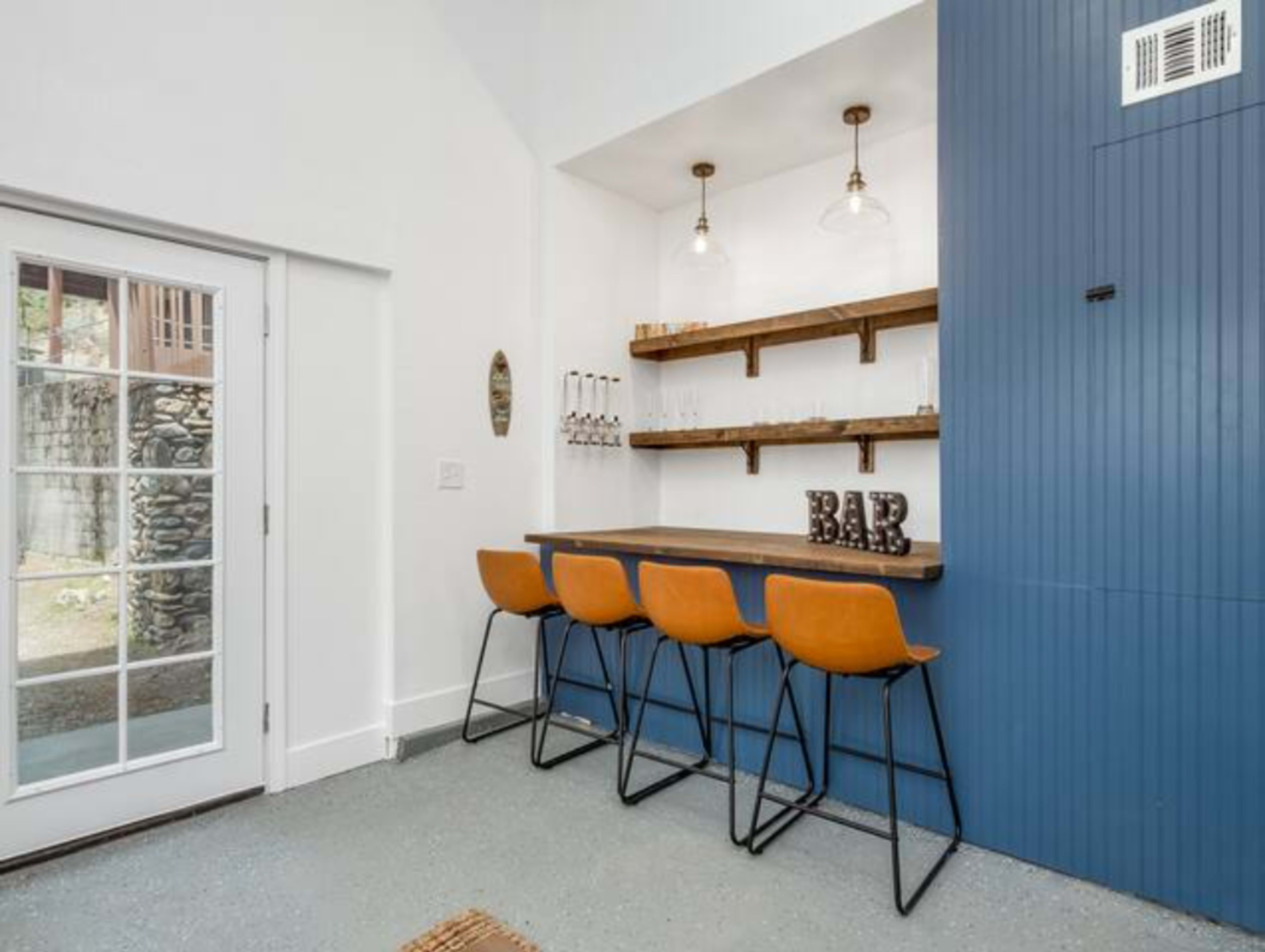 A modern bar area with four orange stools, wooden shelves, and glassware, adjacent to a glass door leading outside.