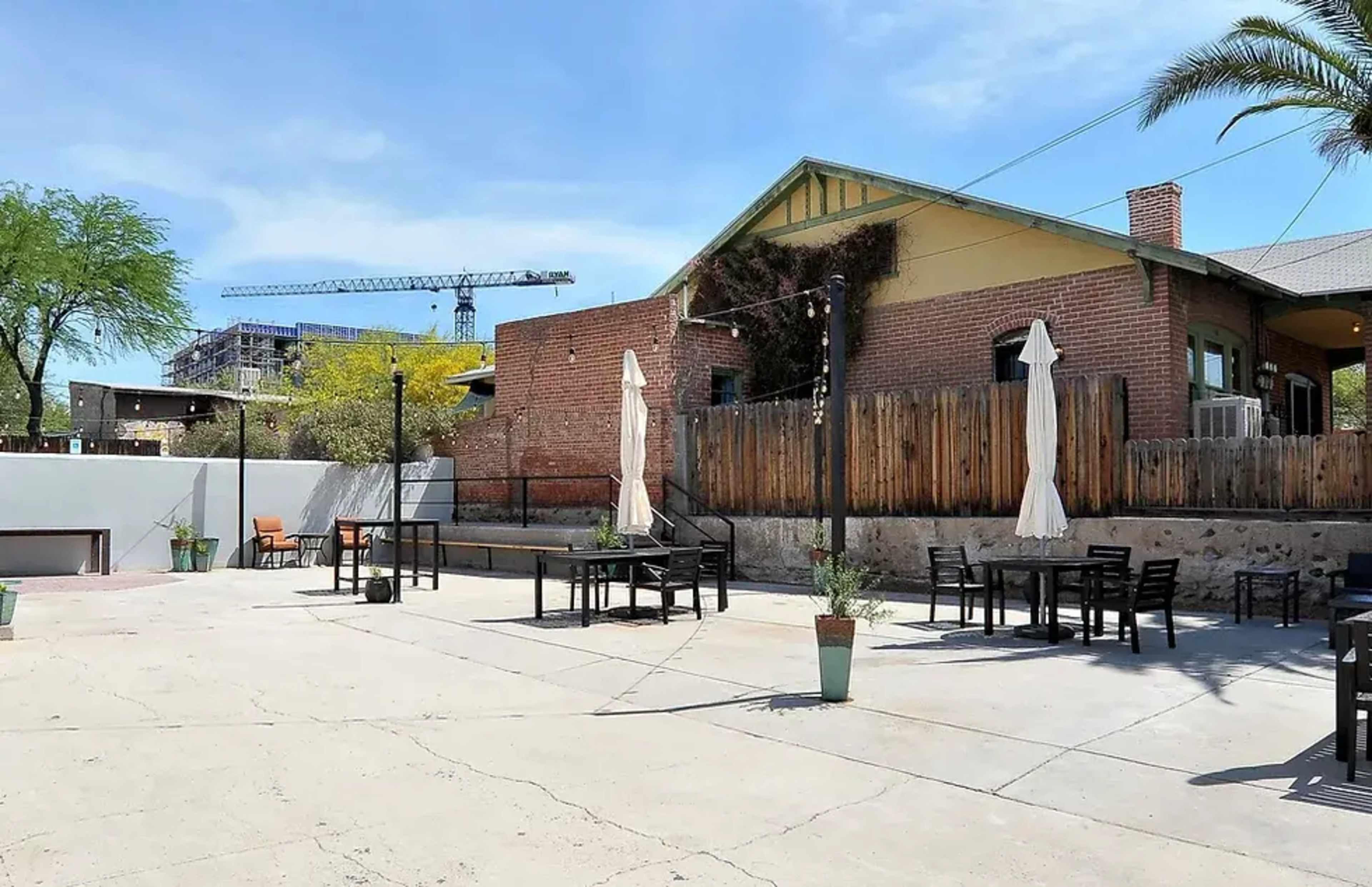 The image shows a spacious outdoor patio area with tables and umbrellas, surrounded by brick buildings and palm trees.