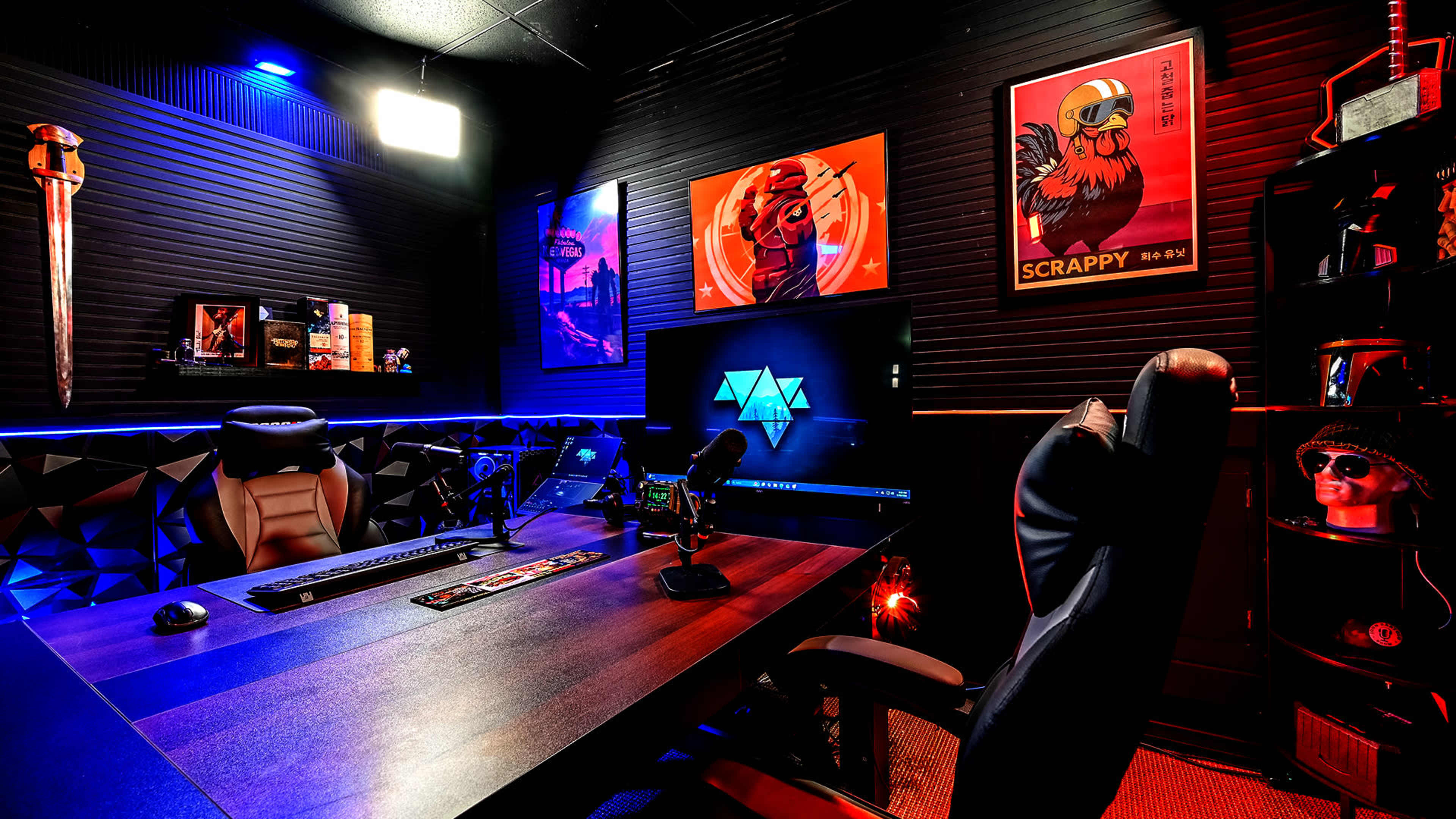 The image shows a modern gaming or streaming setup featuring a dark room with a large desk, multiple microphones, colorful wall art, and ambient lighting.