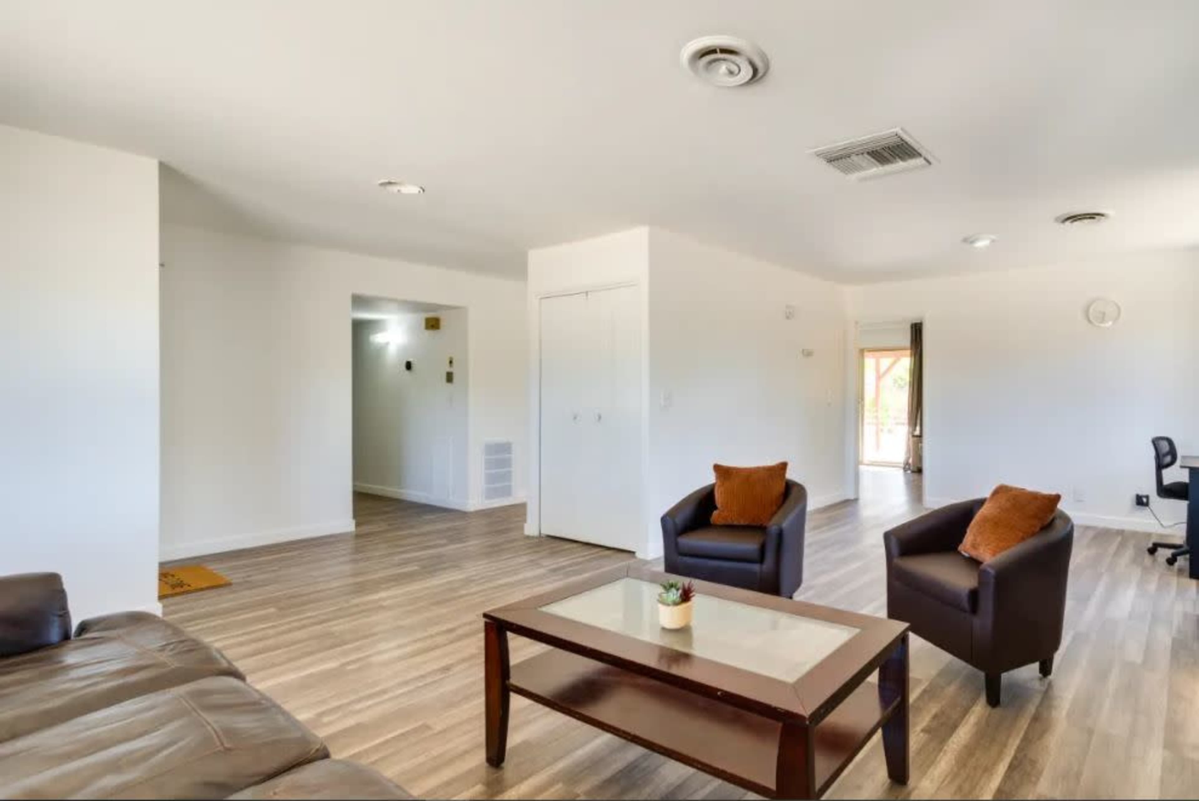 A spacious and well-lit living room with a brown sofa, two black armchairs, and a coffee table, featuring light-colored walls and laminate flooring.