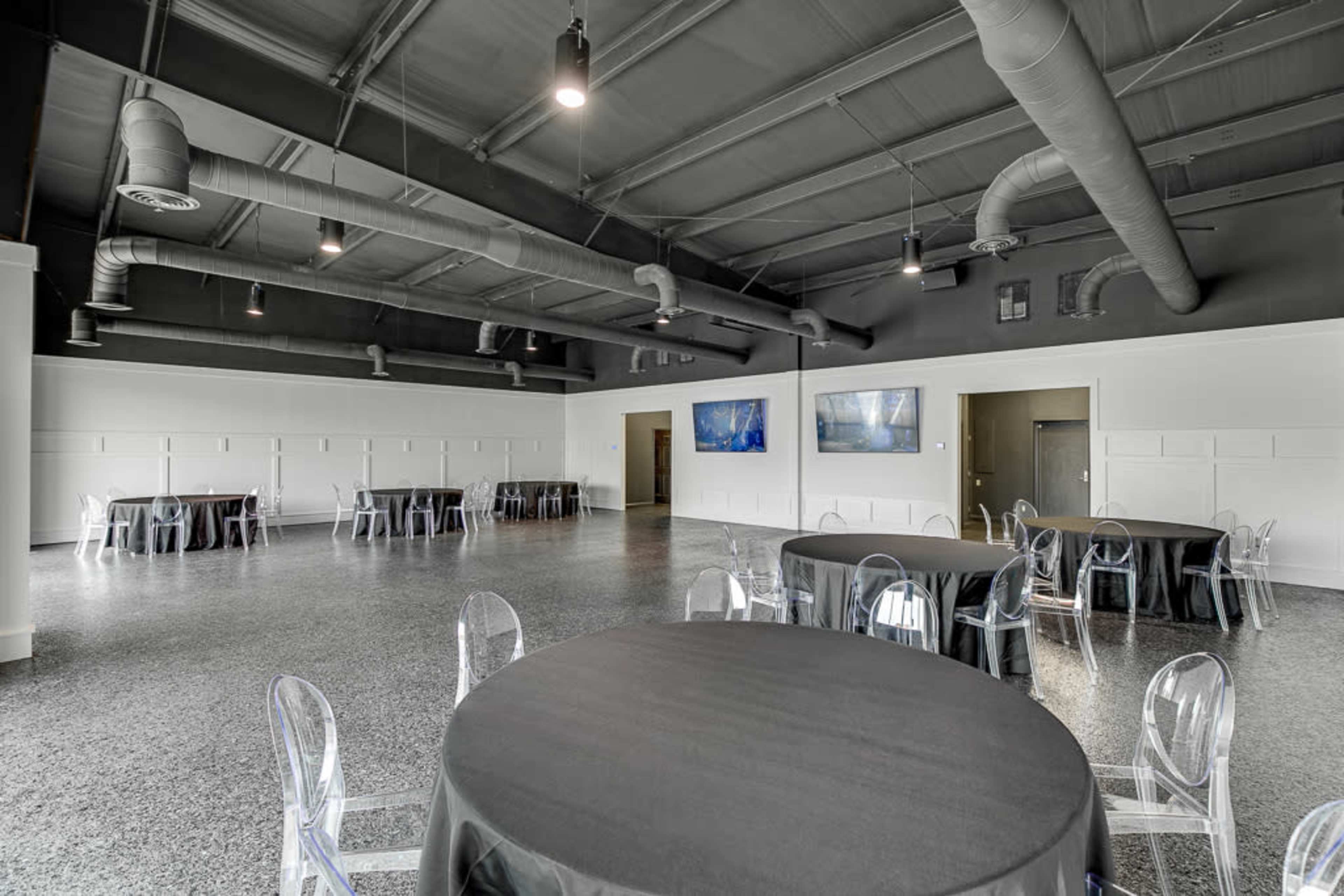 A spacious event room with black round tables covered in dark cloth and clear plastic chairs, featuring gray flooring and two wall-mounted screens.