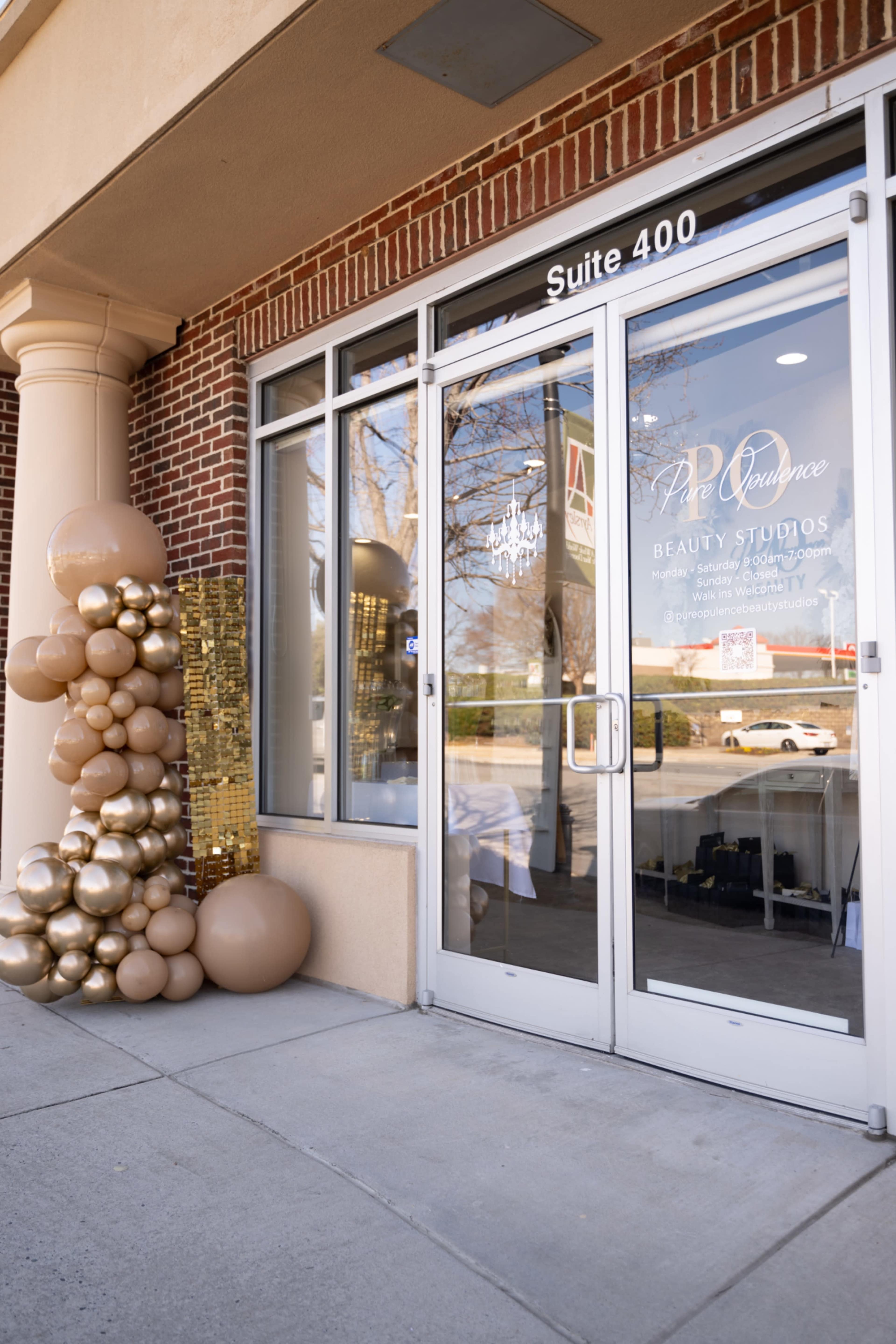 The entrance to a beauty studio features large windows, a modern sign, and a decorative arrangement of gold and beige balloons beside the door.