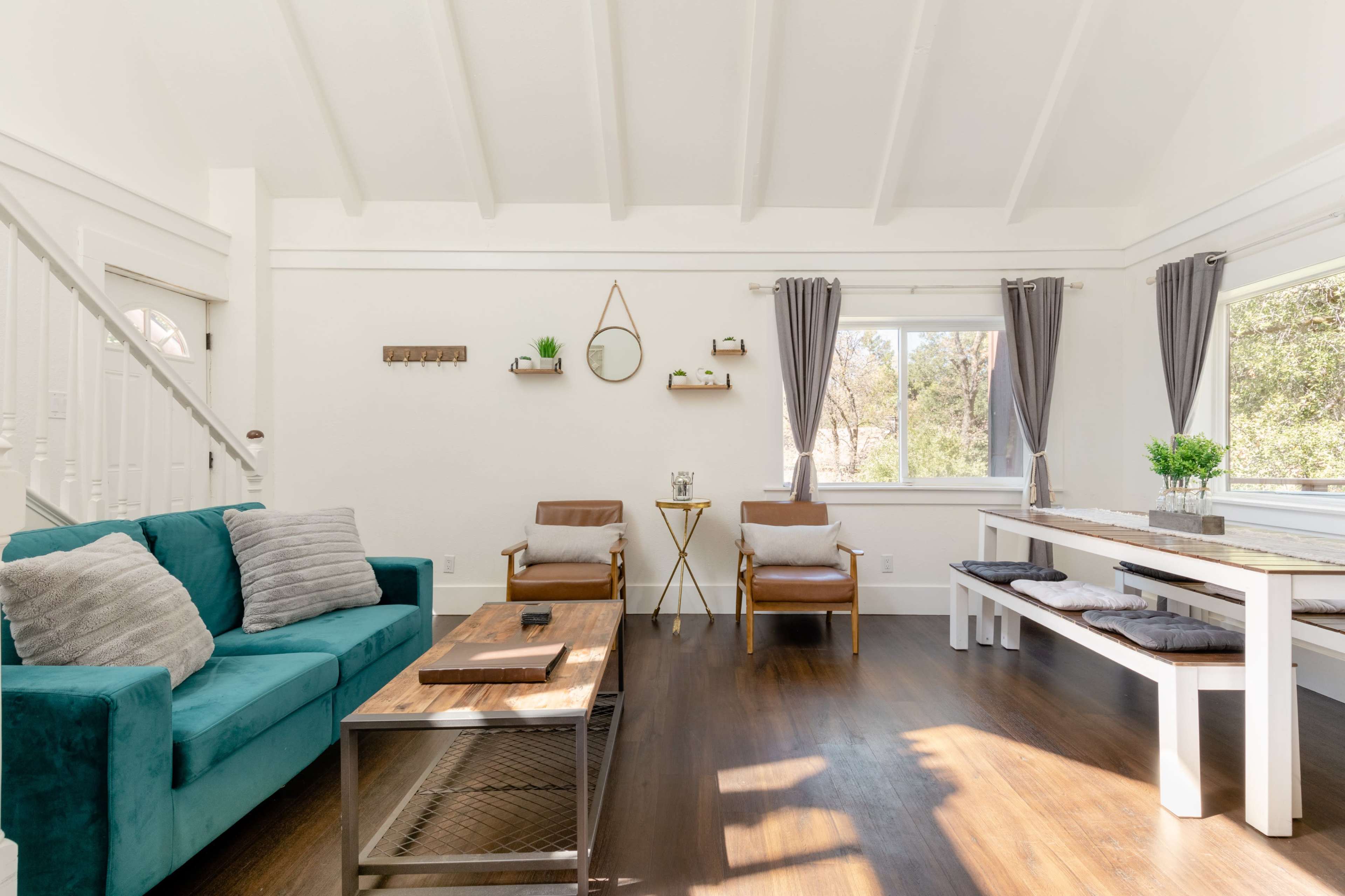 The image shows a bright living room with a teal sofa, two wooden chairs, a coffee table, and a dining table, all situated near large windows.