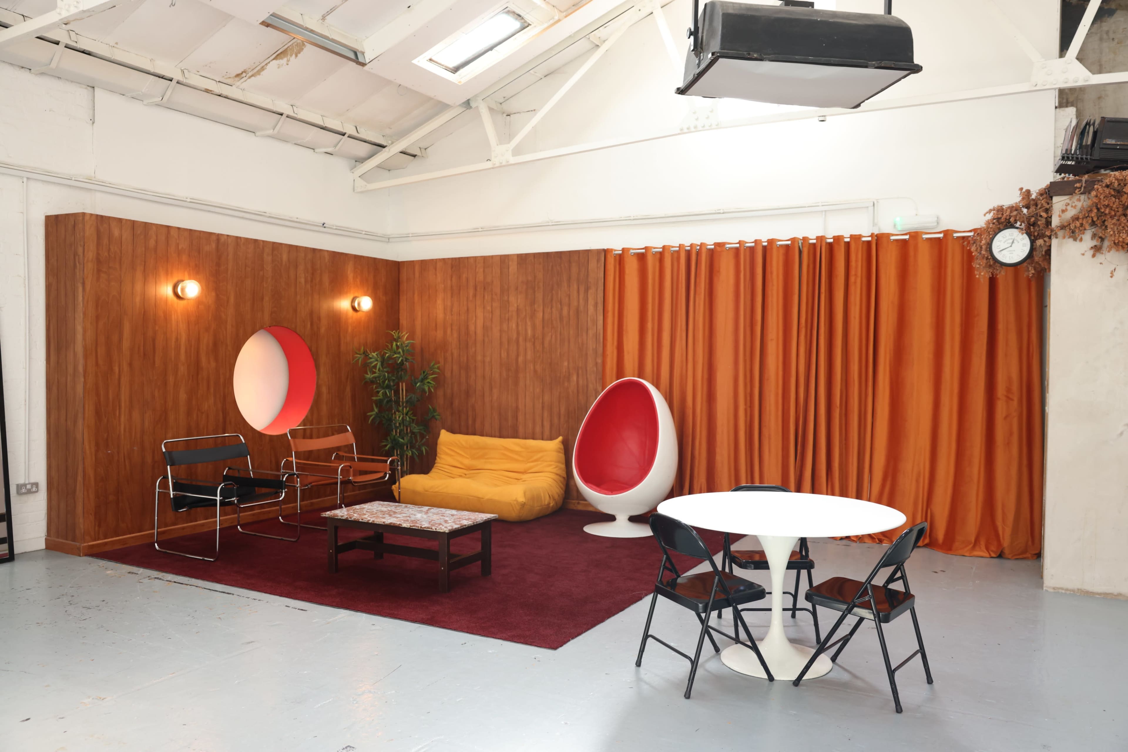 The image shows a modern interior space with a combination of seating options, including a yellow sofa, a red egg chair, and a round table surrounded by black chairs, set against wooden paneling and orange curtains.