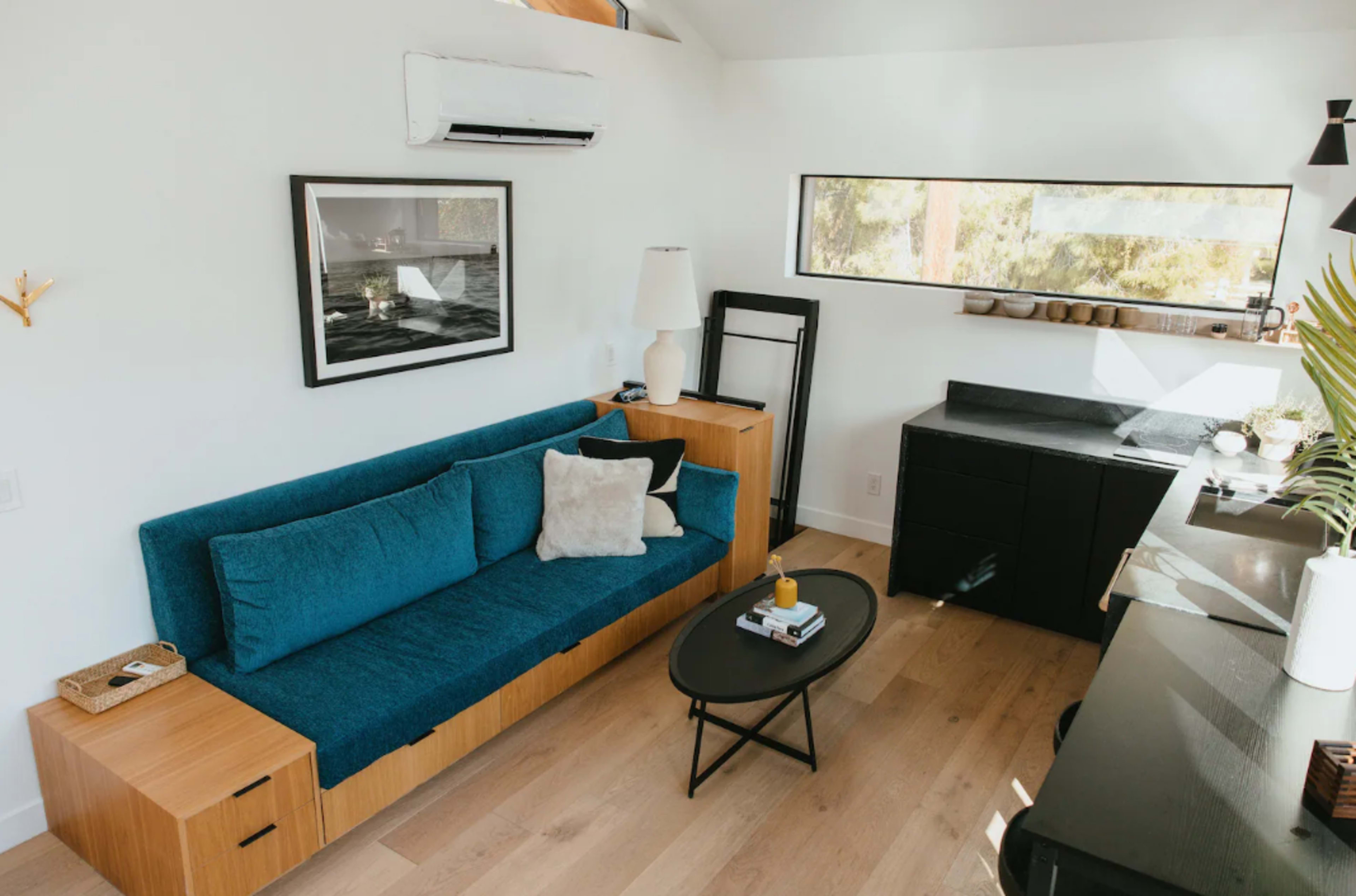 The image shows a modern, minimalist living space featuring a blue sofa, a black coffee table, and a desk against a light-colored wall with a large window.