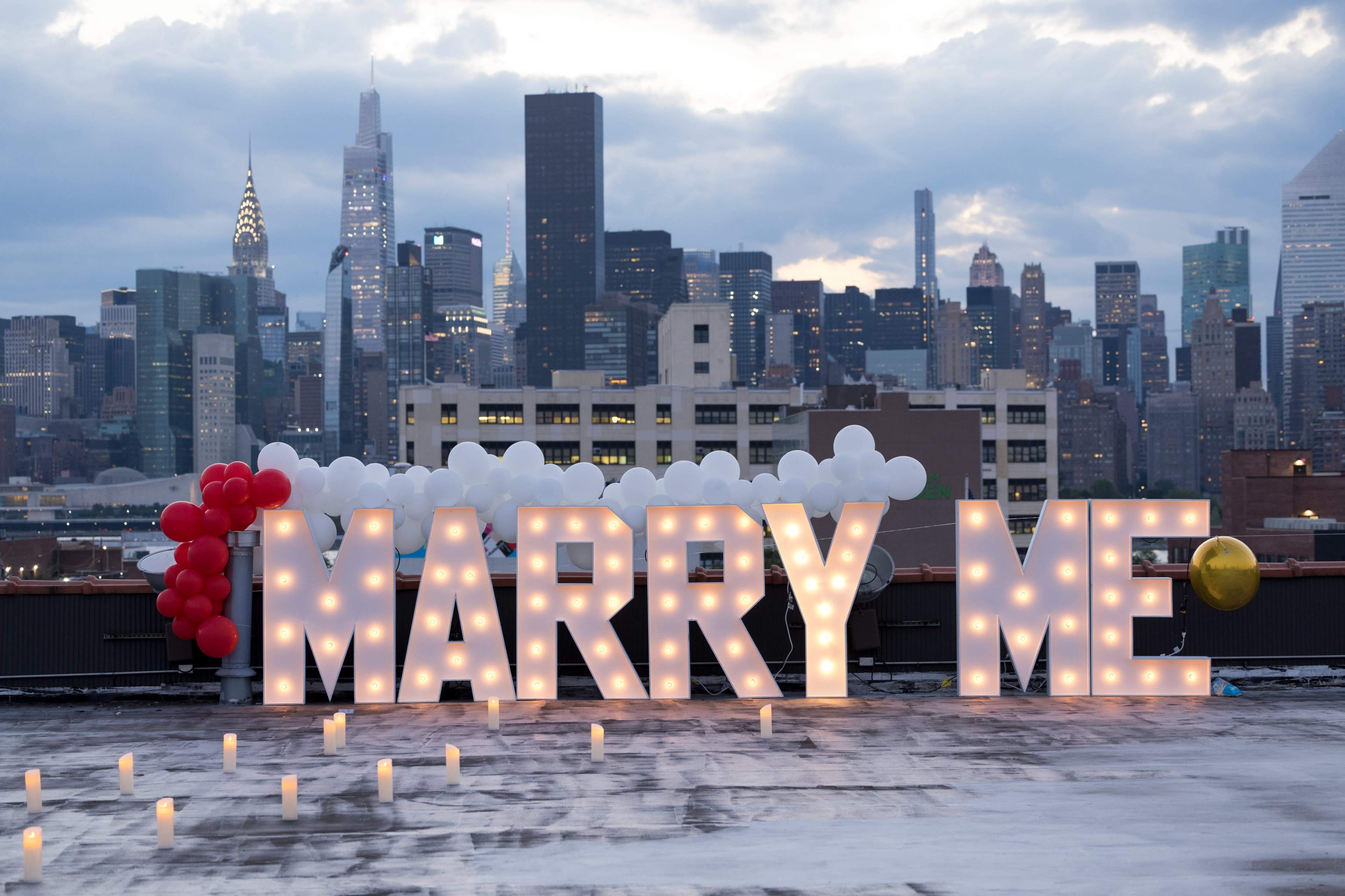 A rooftop proposal scene features large illuminated letters spelling "MARRY ME" against a skyline of New York City.