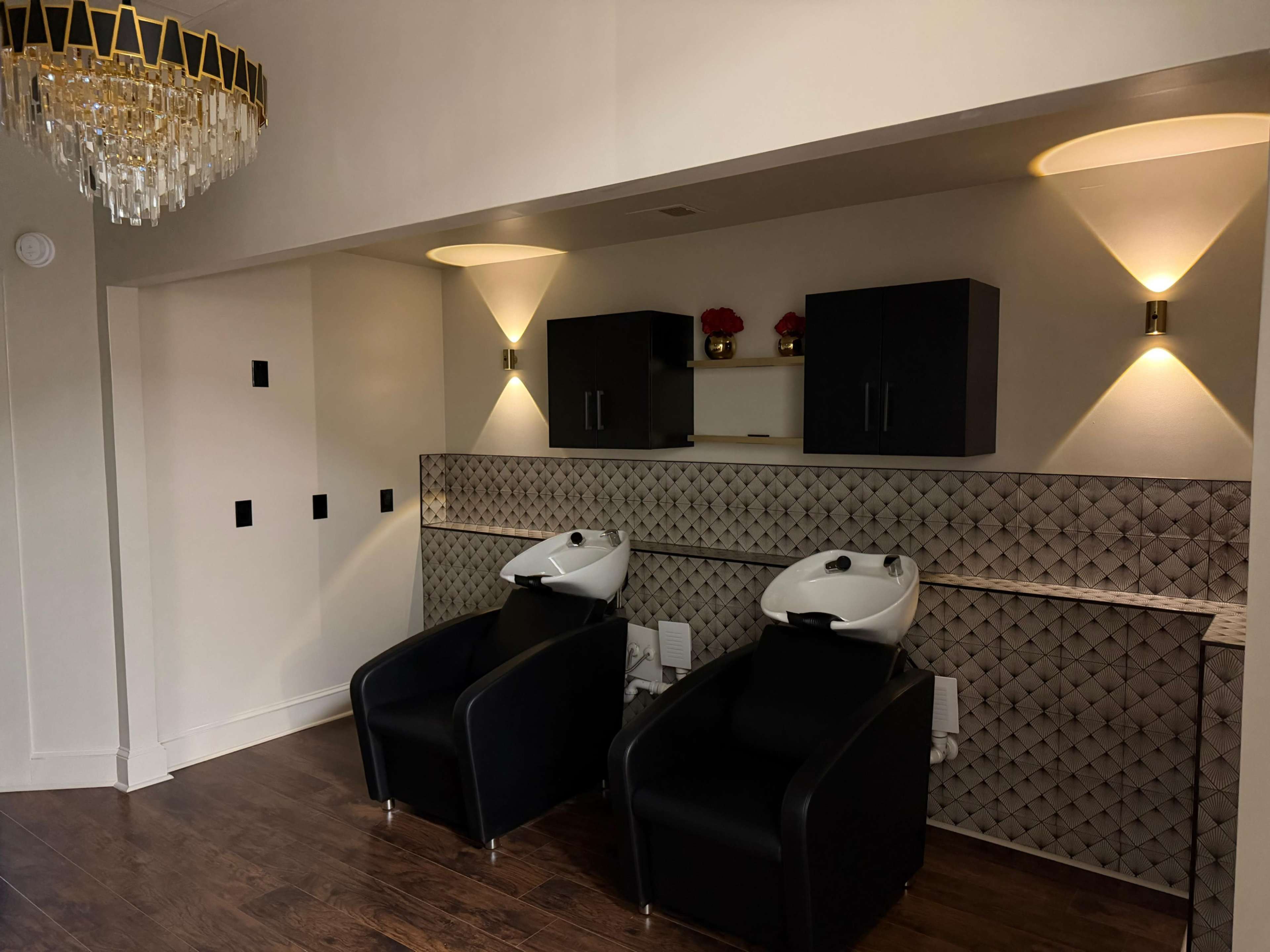 The image shows a salon space with two black shampoo chairs facing a textured wall, illuminated by wall sconces and decorated with cabinets above.