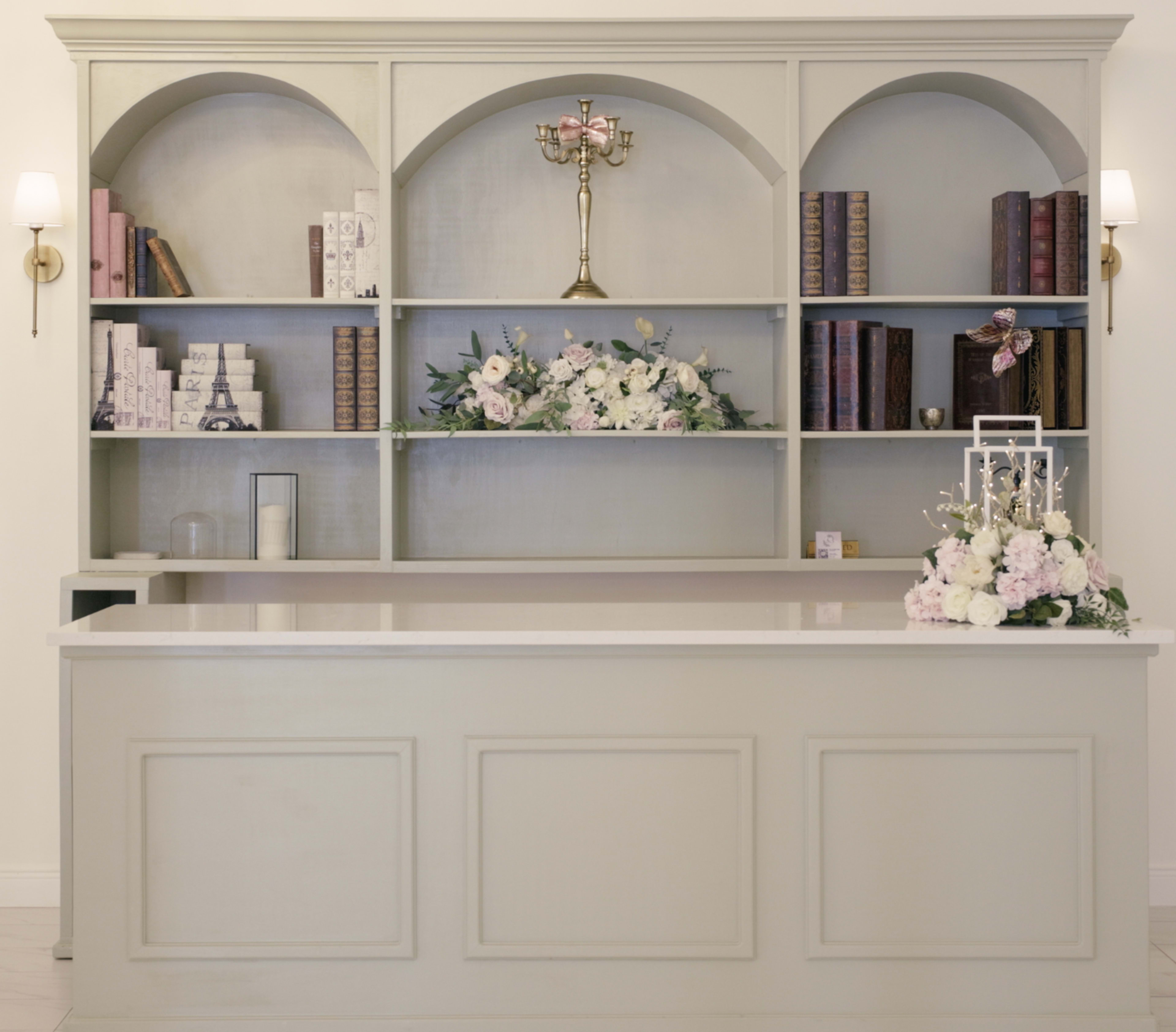 A reception desk is positioned below a decorative shelving unit that displays books, floral arrangements, and a candelabrum.