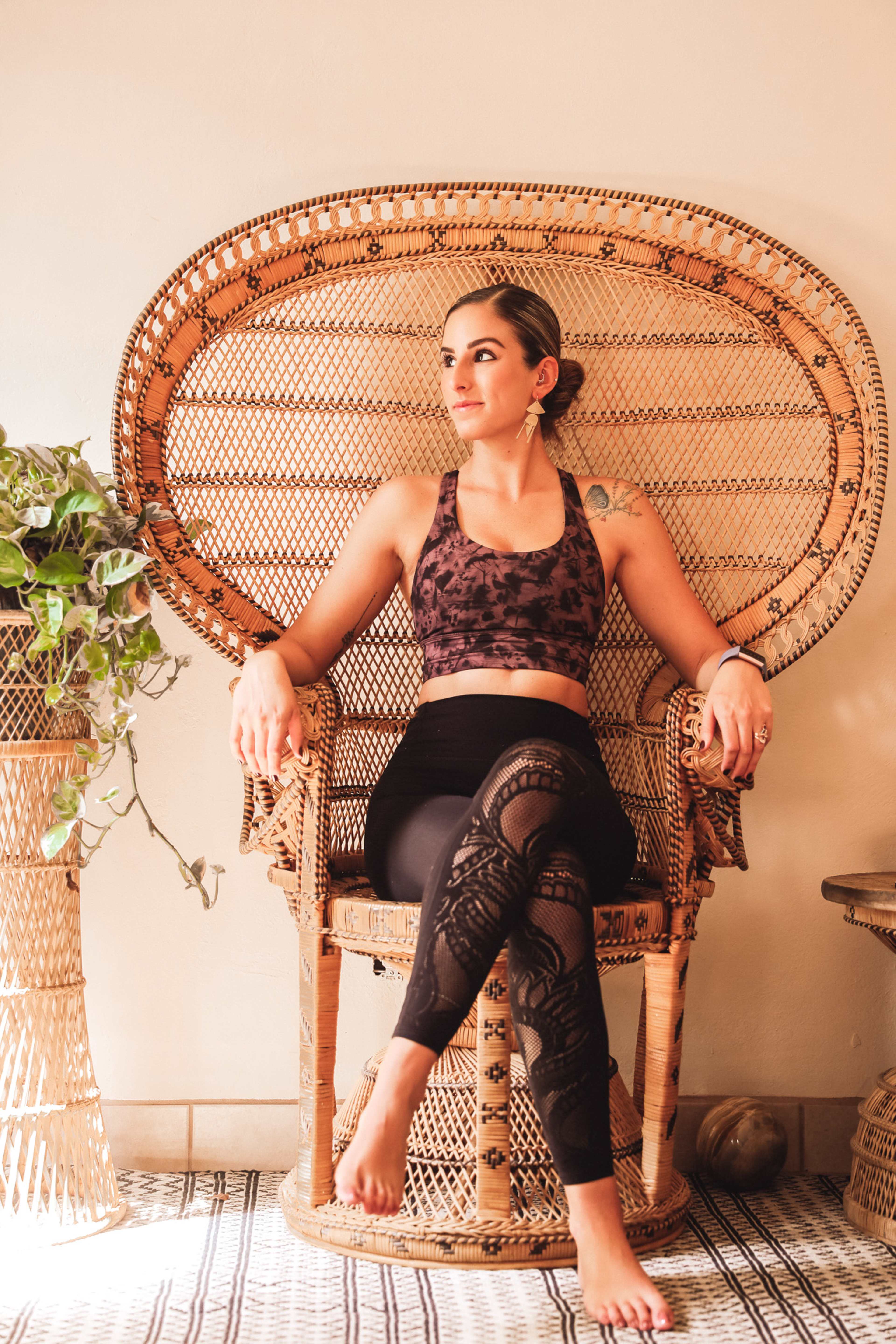 A woman sits in a woven chair, wearing a sports top and patterned leggings, with a plant beside her and a rug underneath.