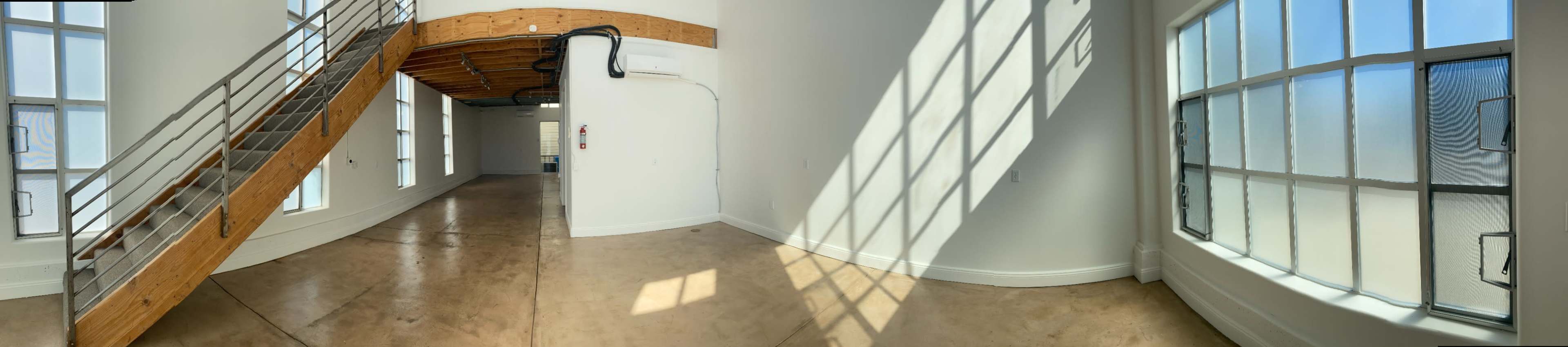 A spacious interior featuring large windows, a lofted area accessible by a wooden staircase, and sunlight casting shadows on the polished concrete floor.