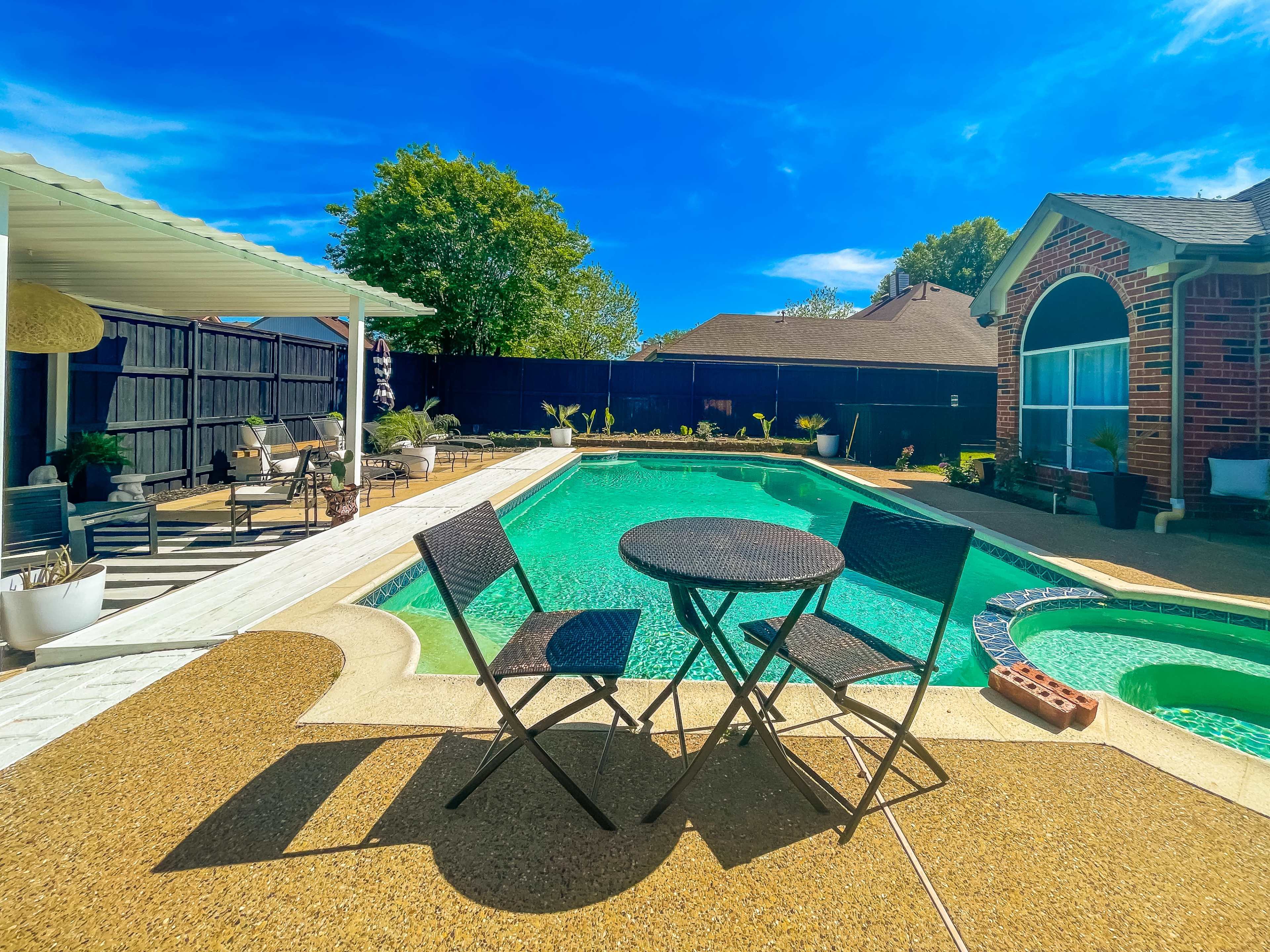 A residential backyard featuring a swimming pool surrounded by concrete, with a table and two chairs set up nearby.