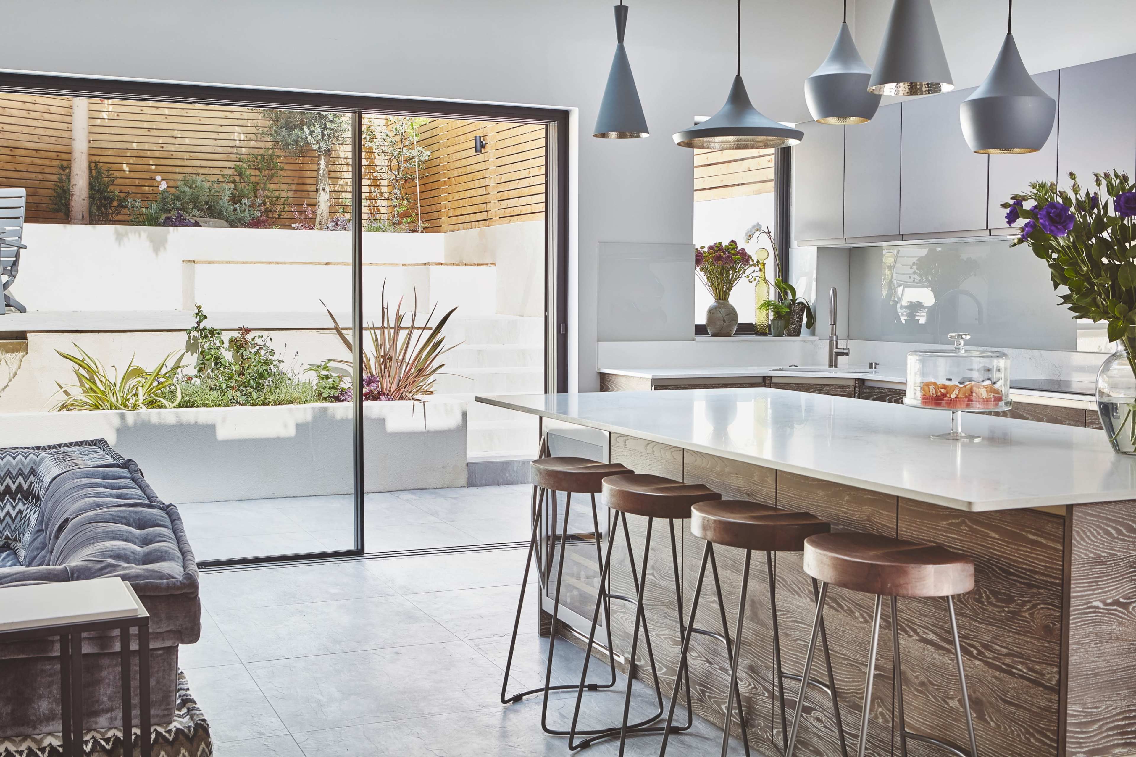 A modern kitchen features a large island with bar stools and an open view to a landscaped outdoor area through sliding glass doors.