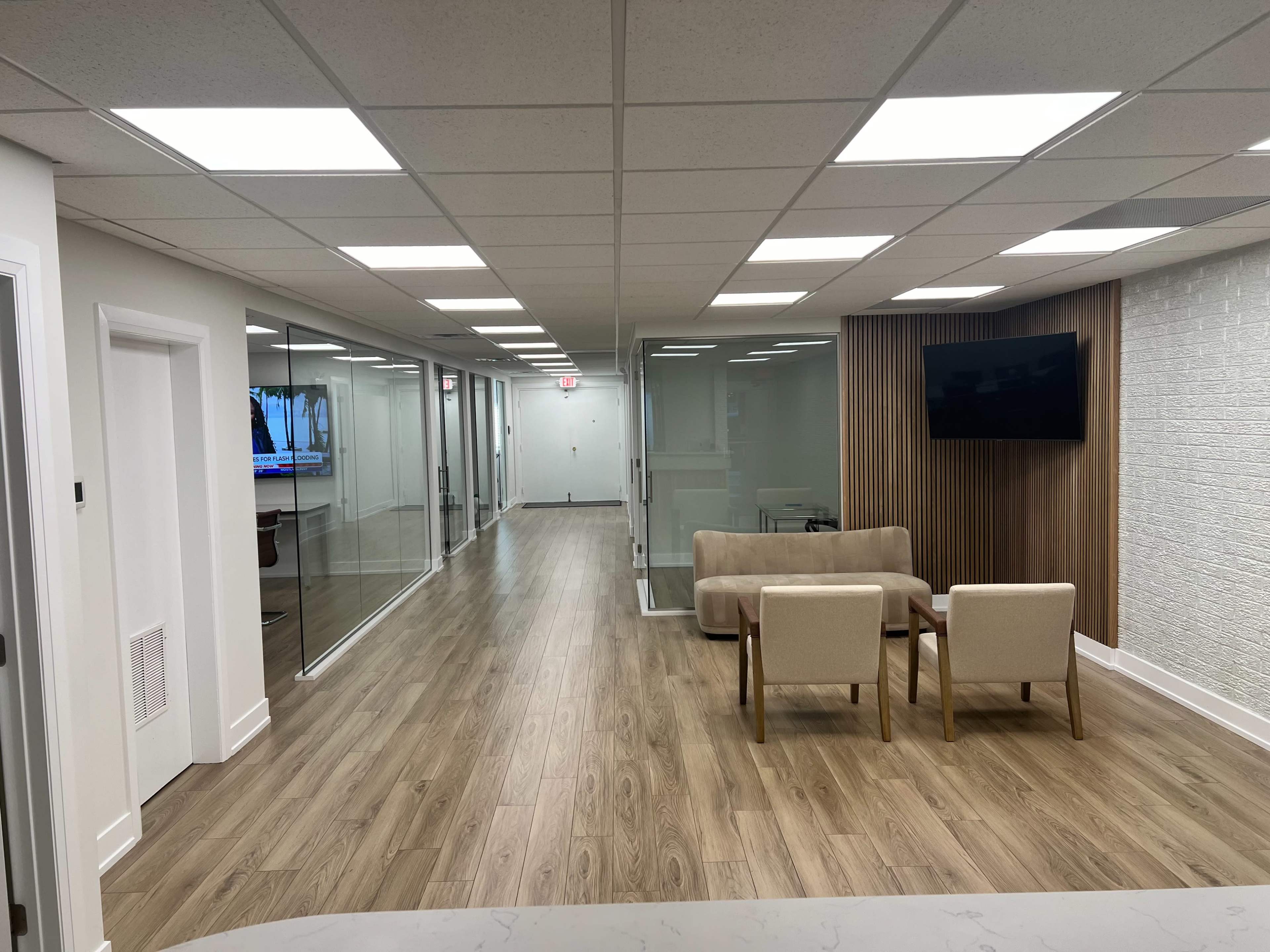 A modern office hallway with glass-walled conference rooms and a seating area featuring two chairs and a sofa.