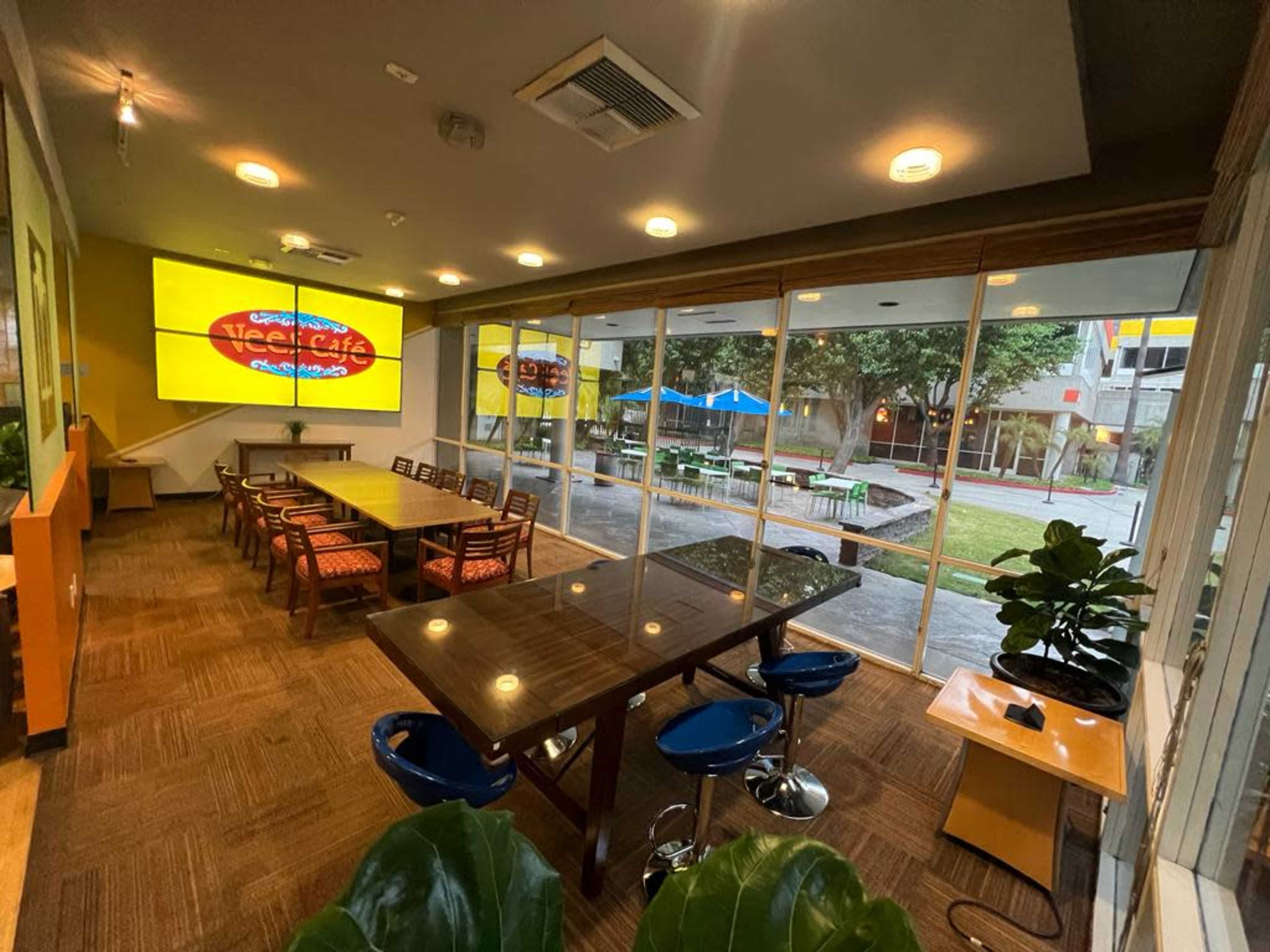 A spacious indoor café area with a large table and several smaller tables, large windows overlooking an outdoor space with seating, and a bright screen displaying the café's name.