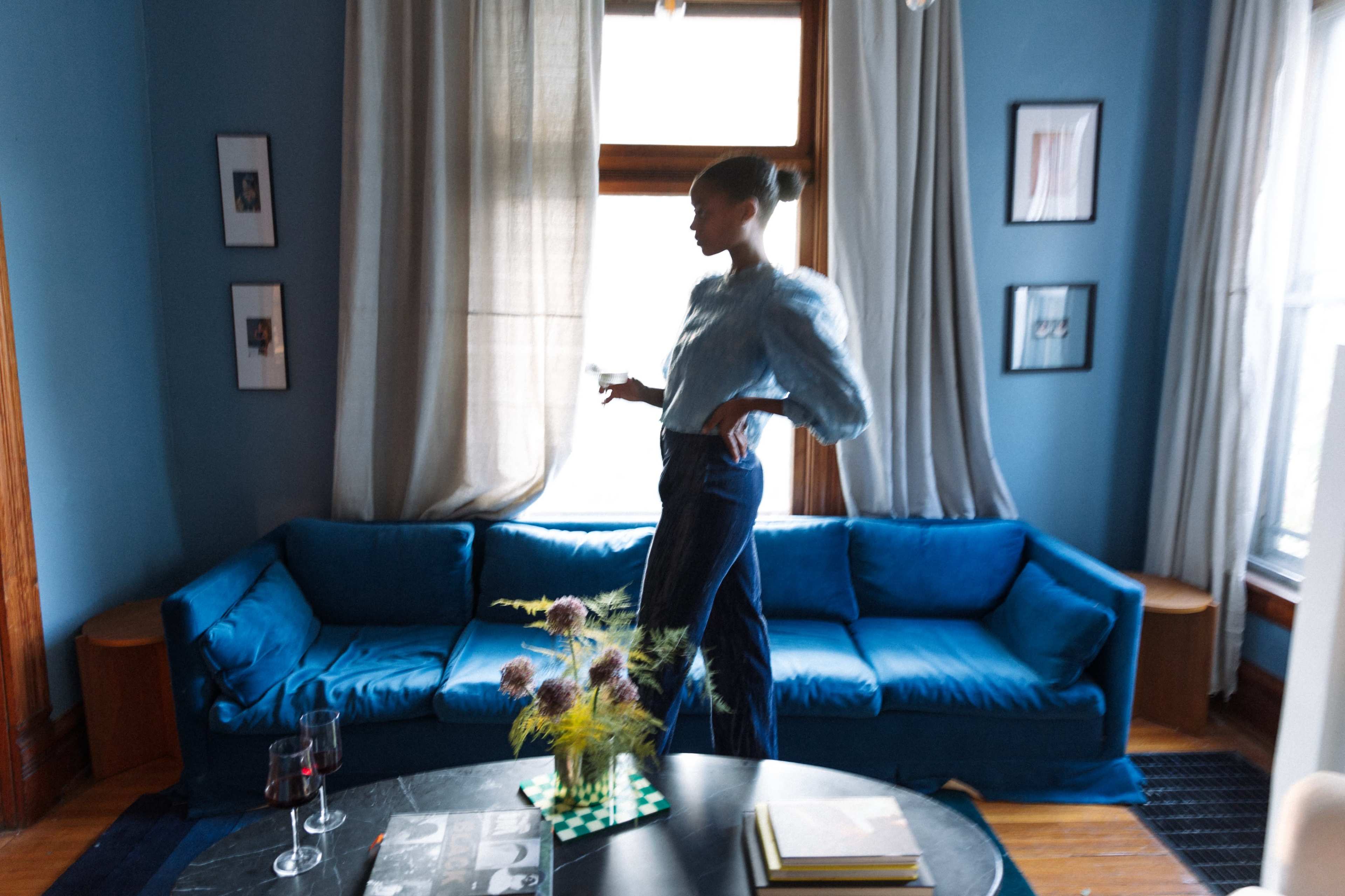 A person stands near a blue sofa in a room with blue walls, holding a drink and surrounded by various decorative elements.