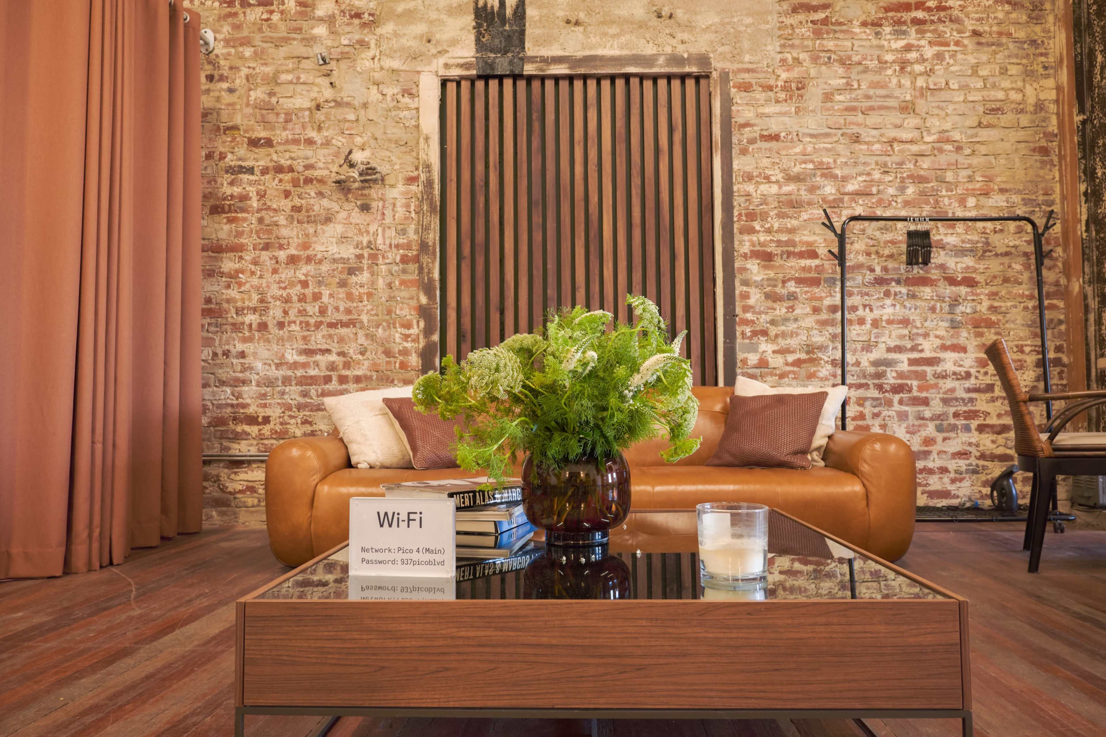 A modern living room features a brown leather sofa, a wooden coffee table with a candle and books, and exposed brick walls.