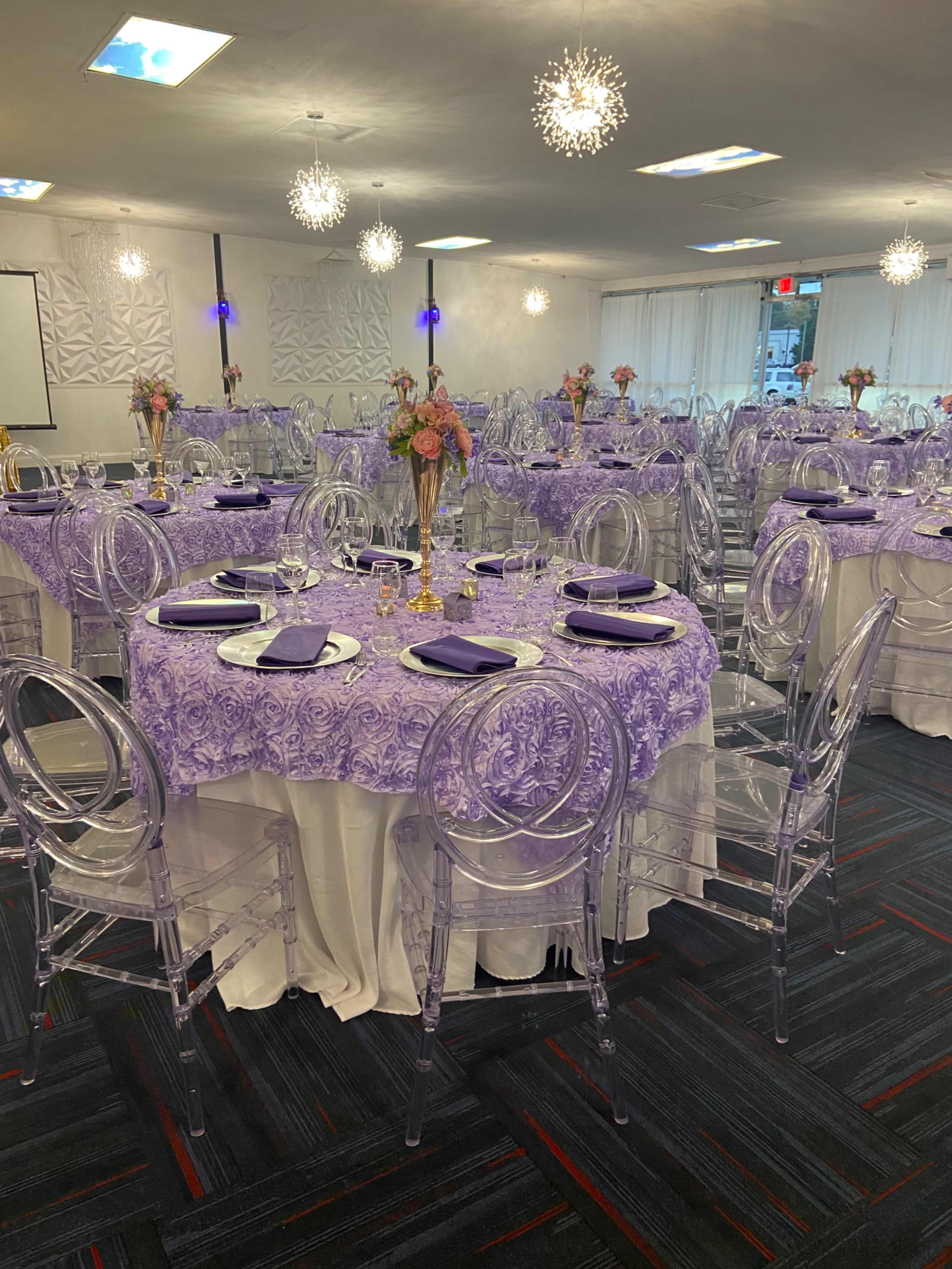 The image shows a banquet hall set for an event, featuring tables covered with purple lace tablecloths and clear acrylic chairs.
