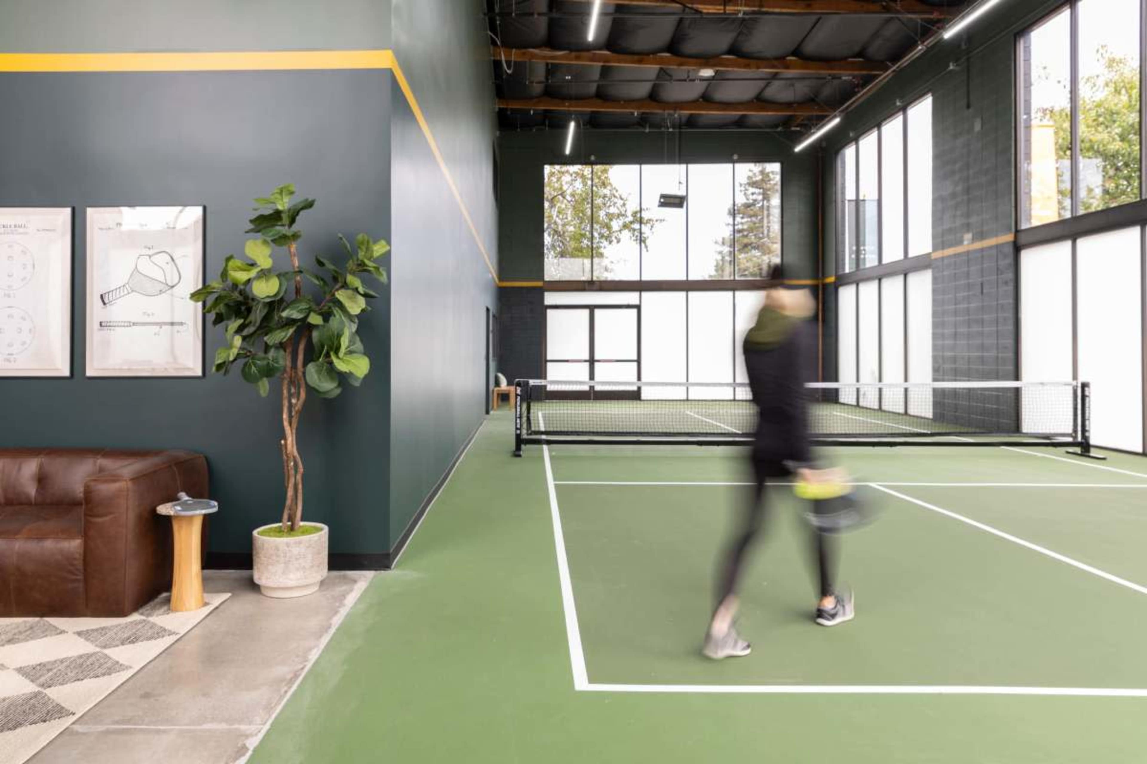 The image shows an indoor tennis court with large windows, a green playing surface, a sofa, a small table, and a potted plant in the corner.