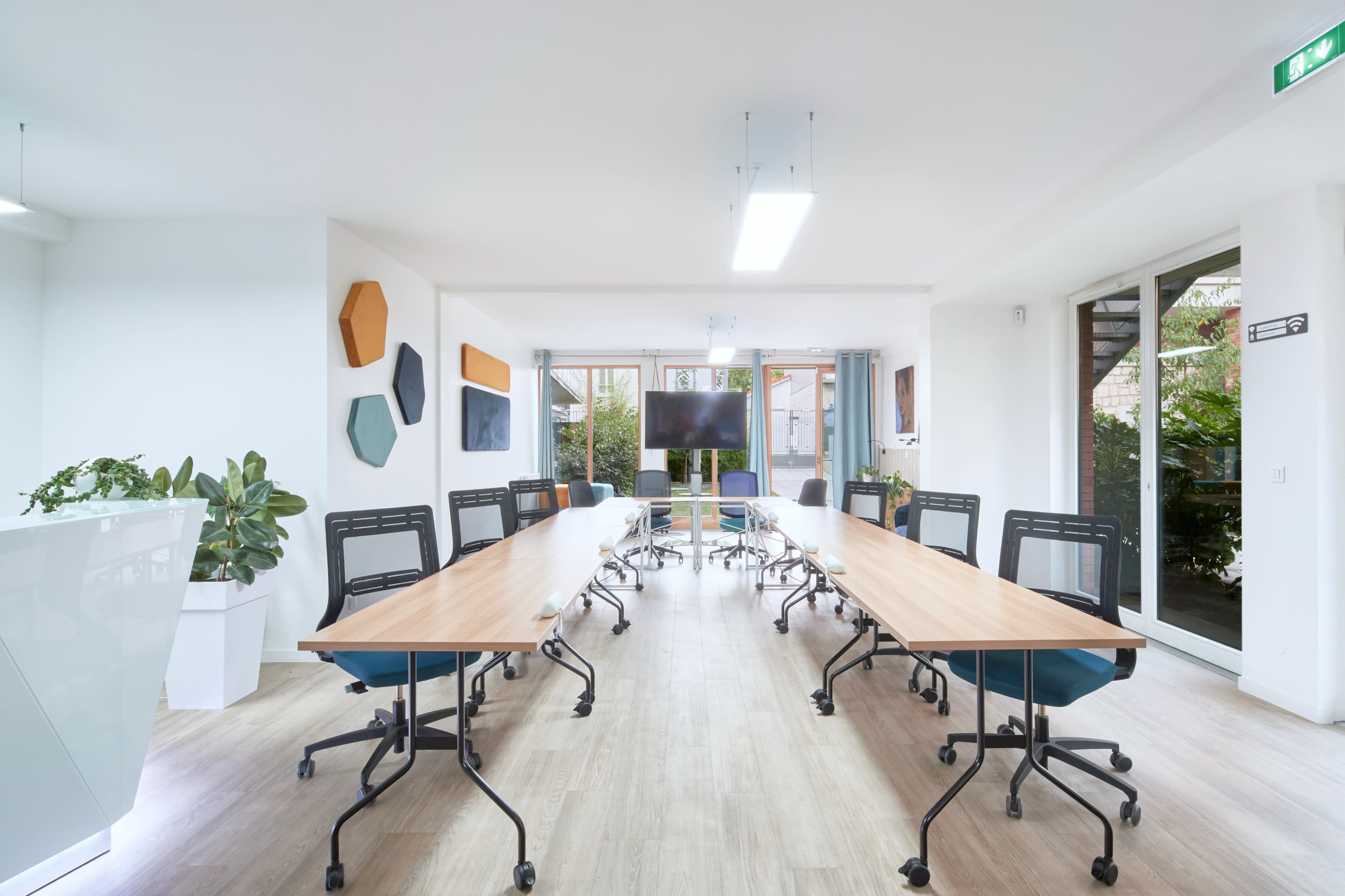 A modern conference room features a long wooden table surrounded by mesh chairs, with large windows providing natural light and a view of greenery outside.