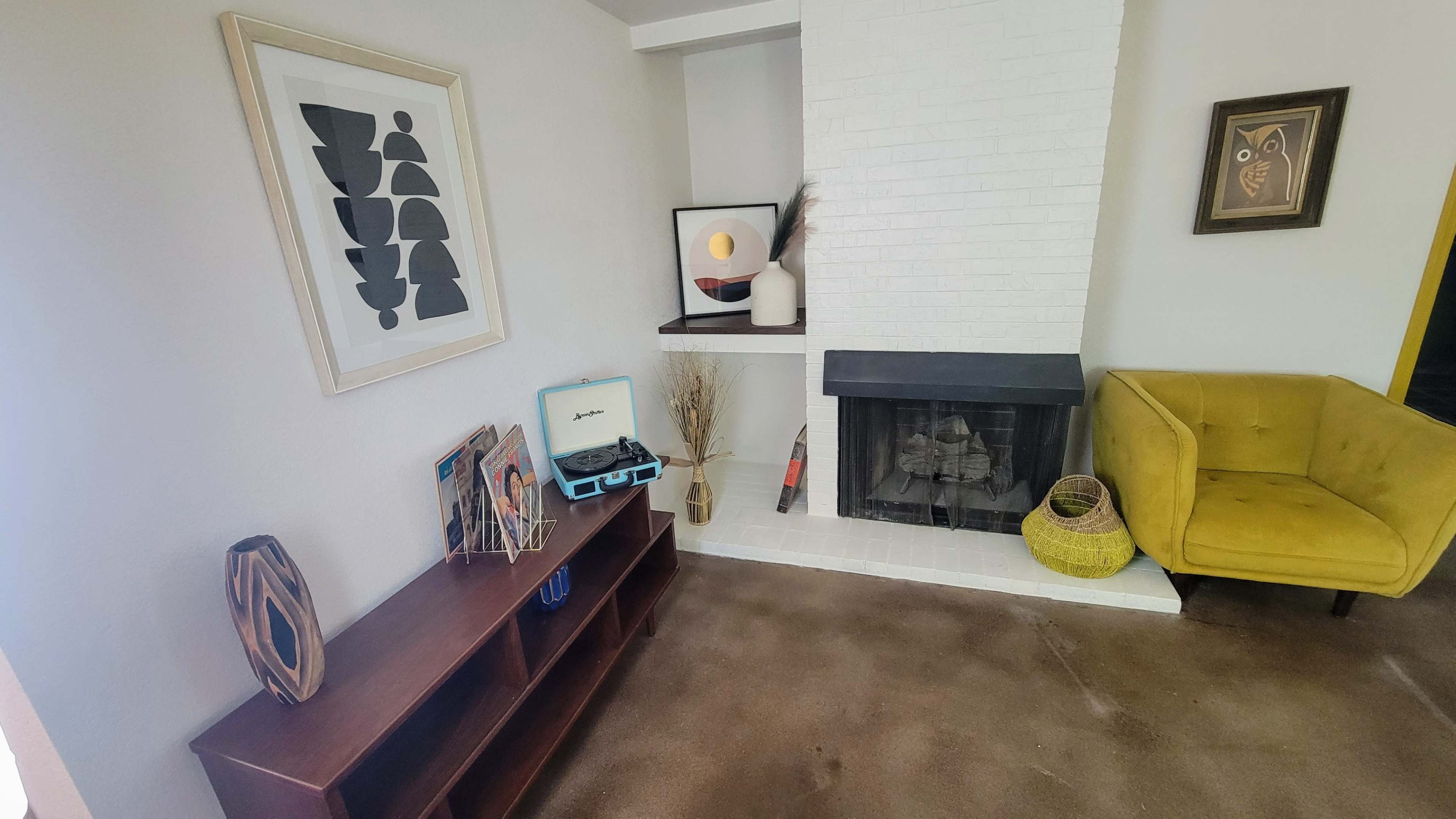 A living room with a yellow chair, a white brick fireplace, a wooden shelf holding magazines, and decorative items on the mantel and nearby.