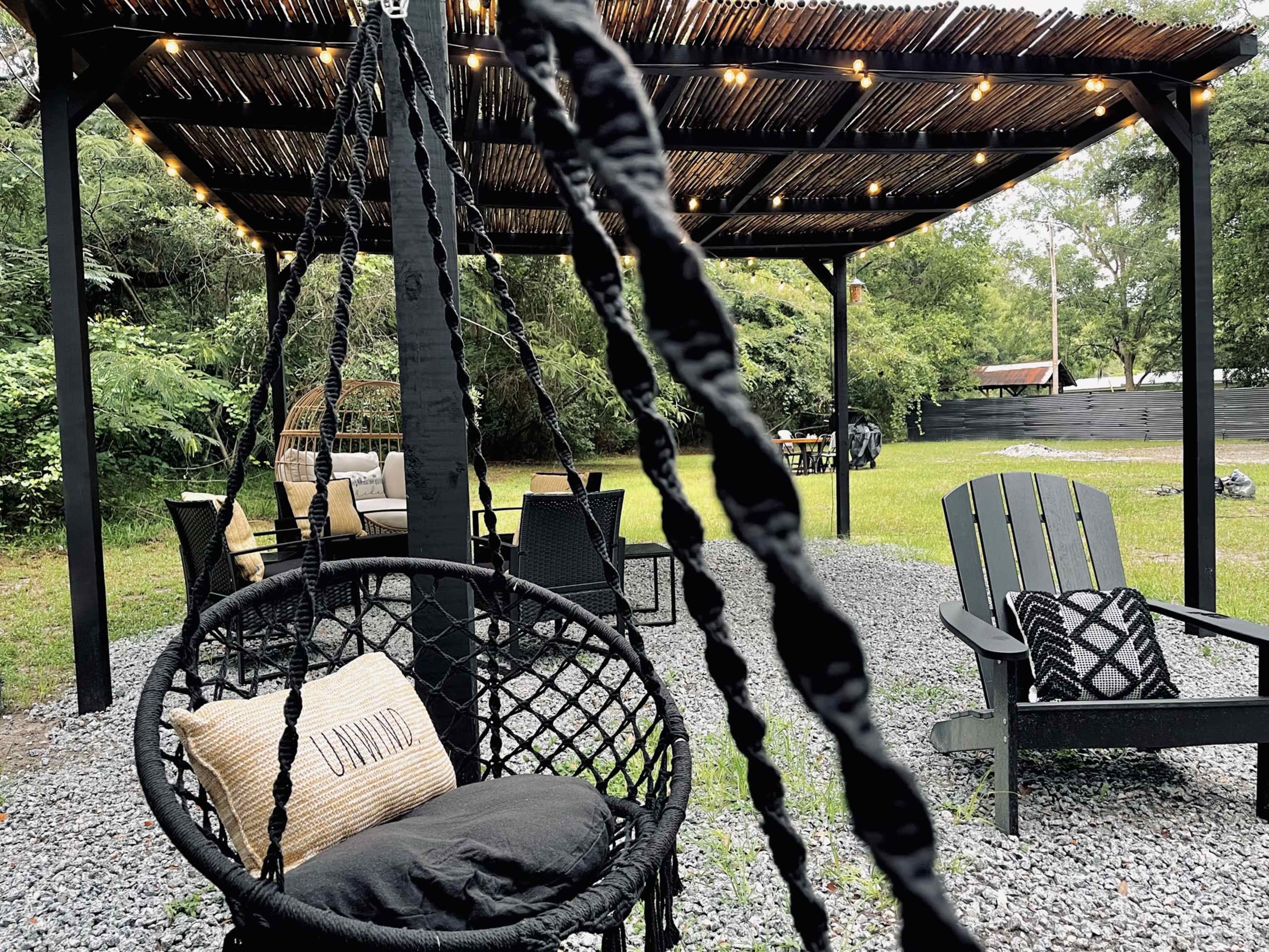 A shaded outdoor seating area features hanging chairs, a cozy couch, and string lights above, surrounded by greenery and gravel.