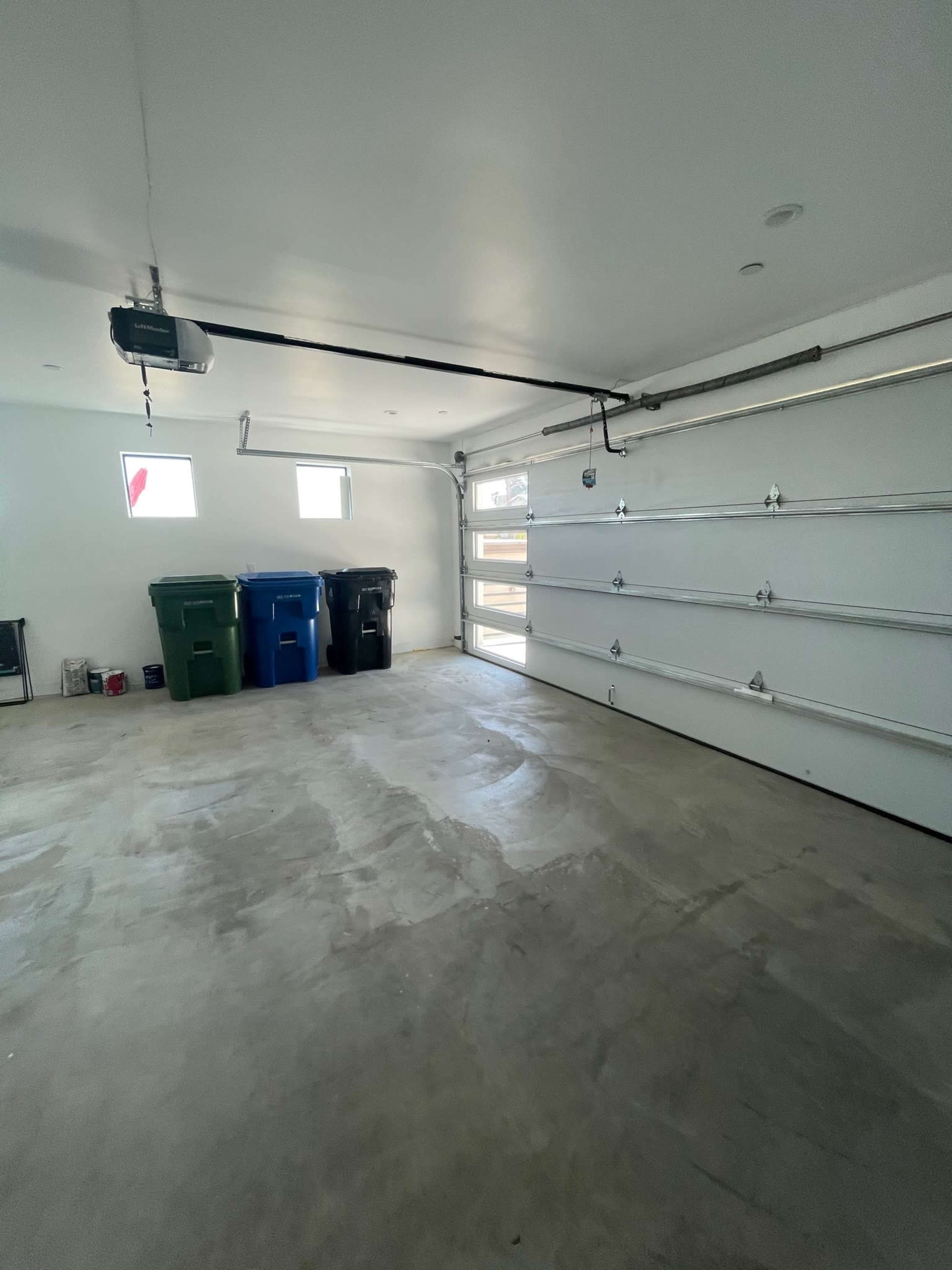 The image shows an empty garage with a concrete floor, two windows, and three color-coded trash bins along one wall.