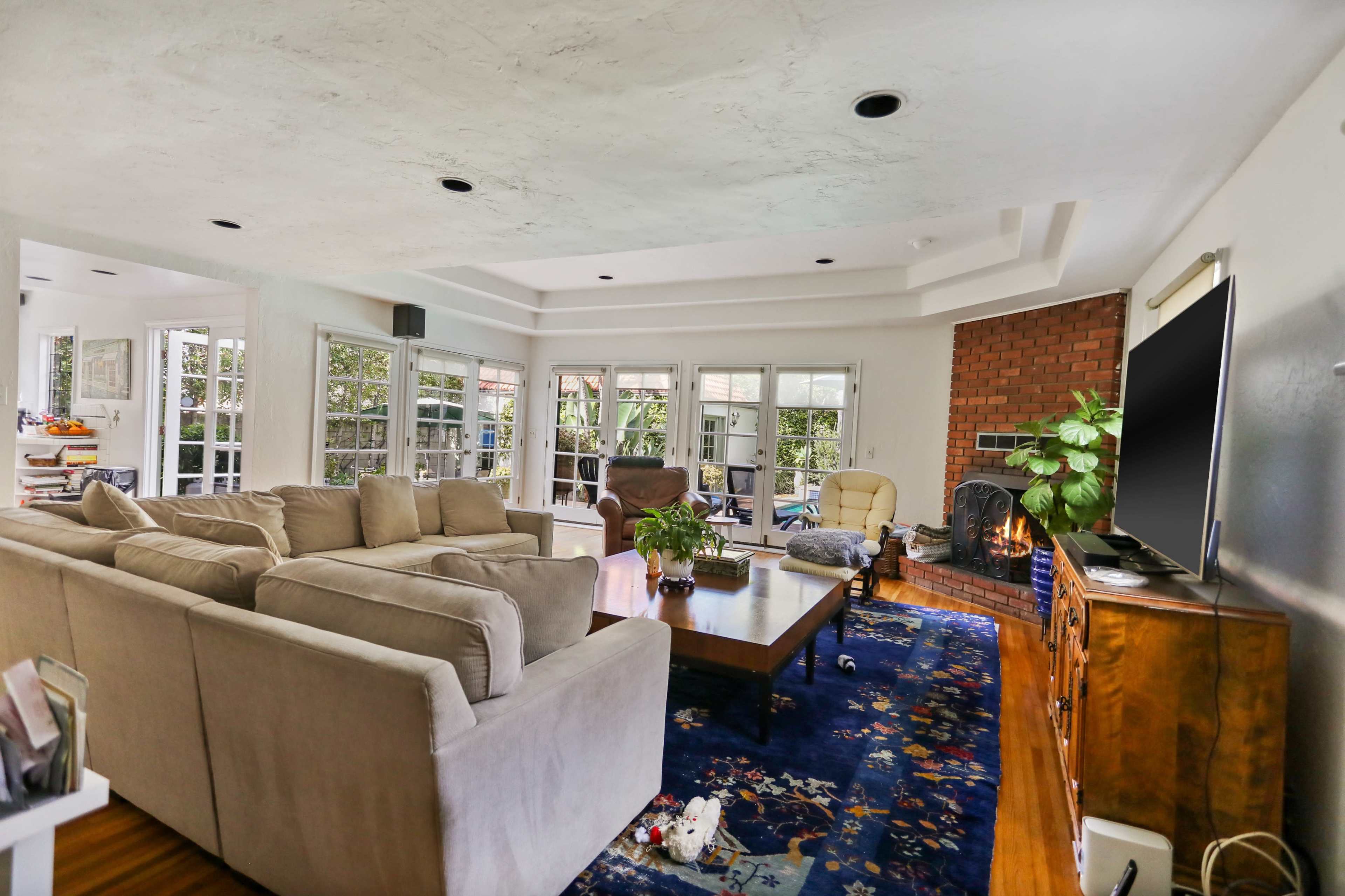 A spacious living room features a sofa set, a wooden coffee table, and a brick fireplace, with large windows providing views of the outdoors.