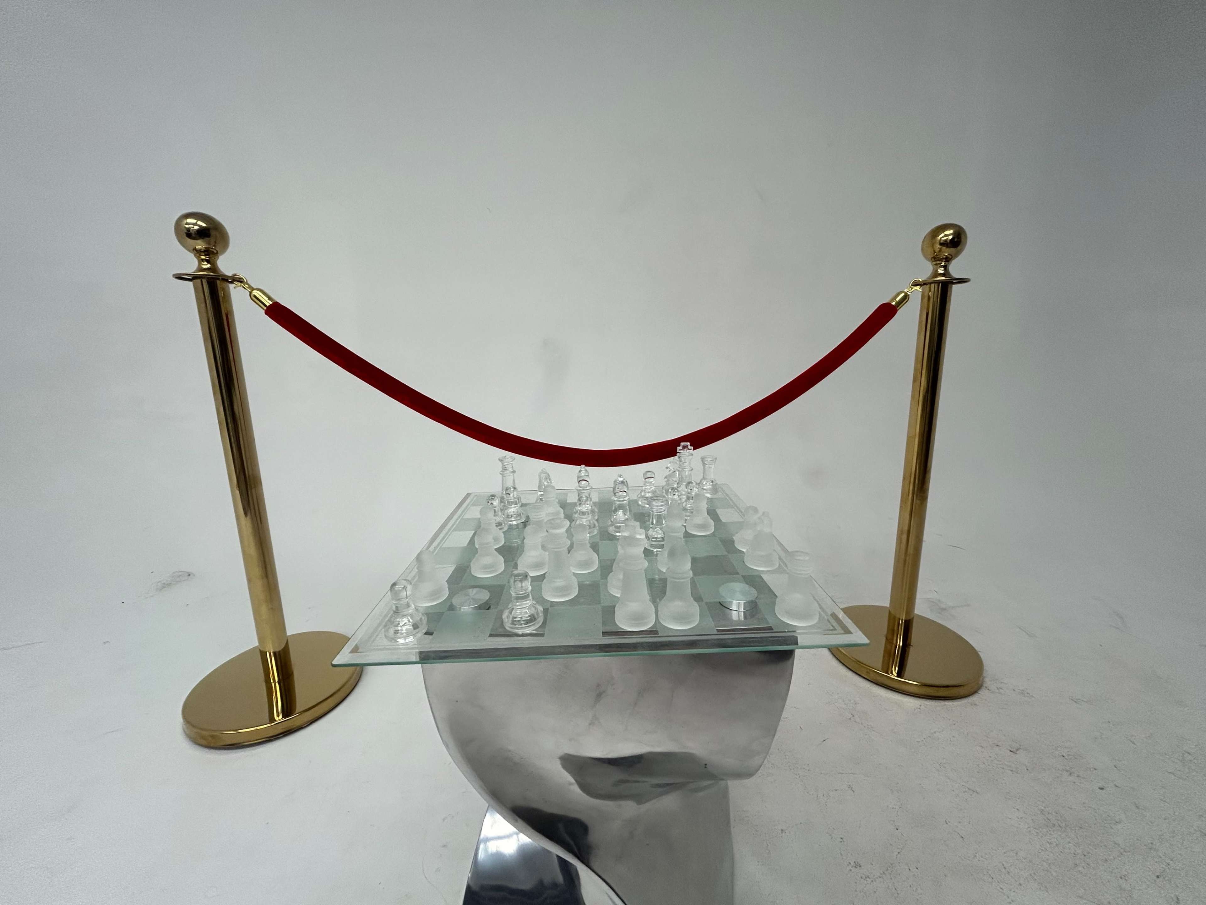 A glass chessboard with clear pieces is placed on top of a reflective, metallic head sculpture, surrounded by a red velvet rope and golden stanchions.