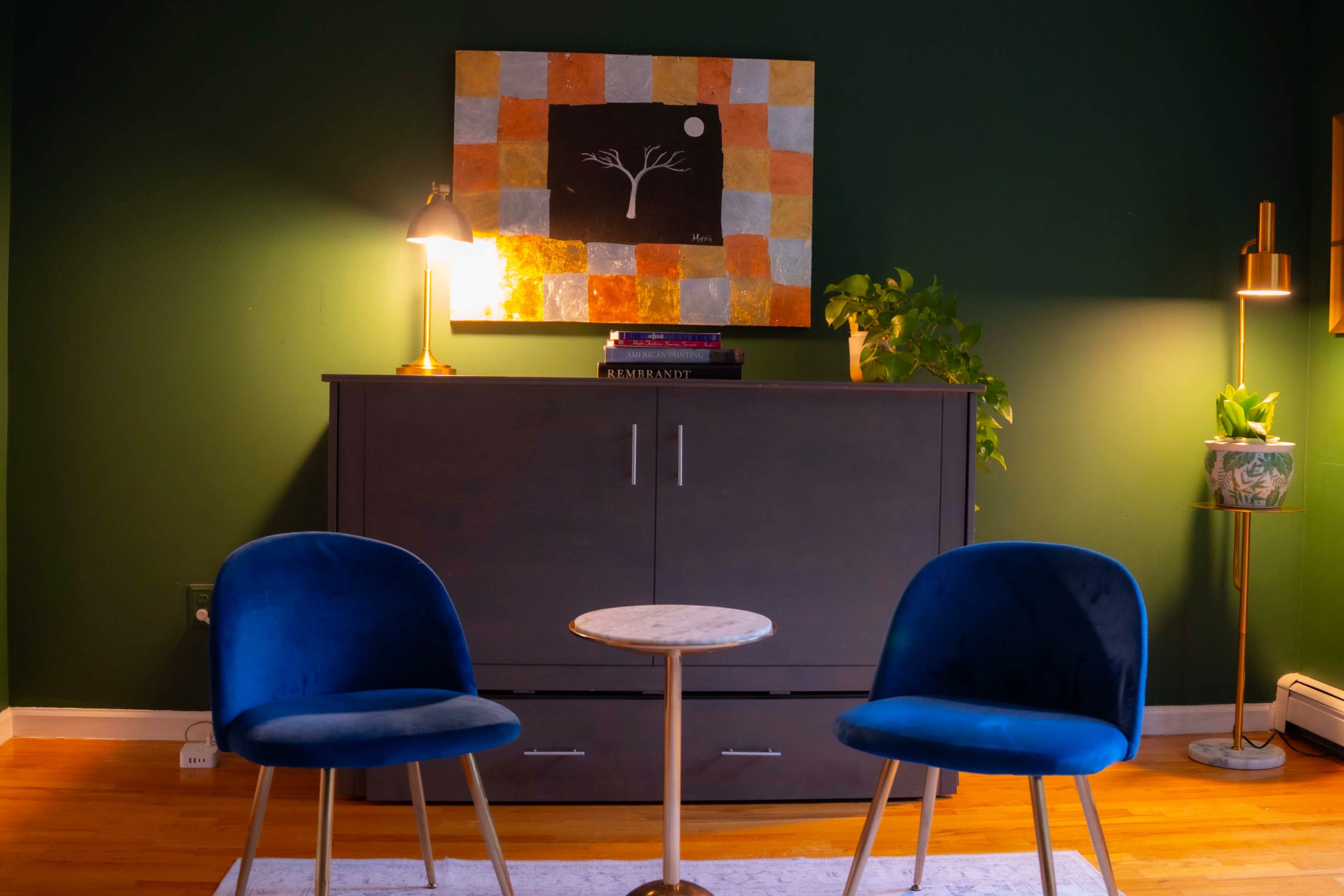 The image shows a cozy living room with two blue chairs facing a small round table, a dark cabinet underneath a colorful painting, and a lamp illuminating the space.