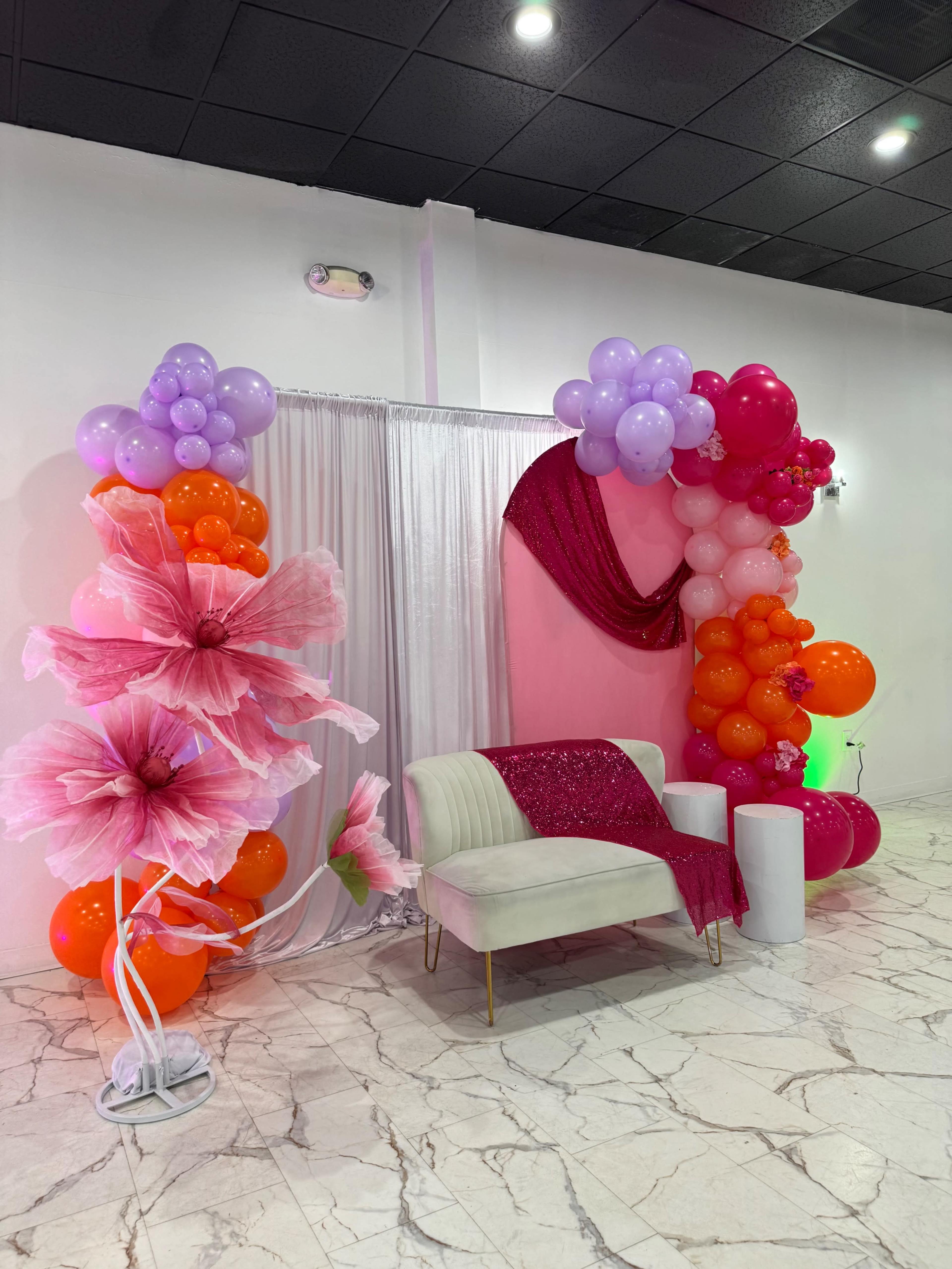 The image depicts a decorated event space featuring a white couch, colorful floral and balloon arrangements, and a backdrop of draped white fabric.