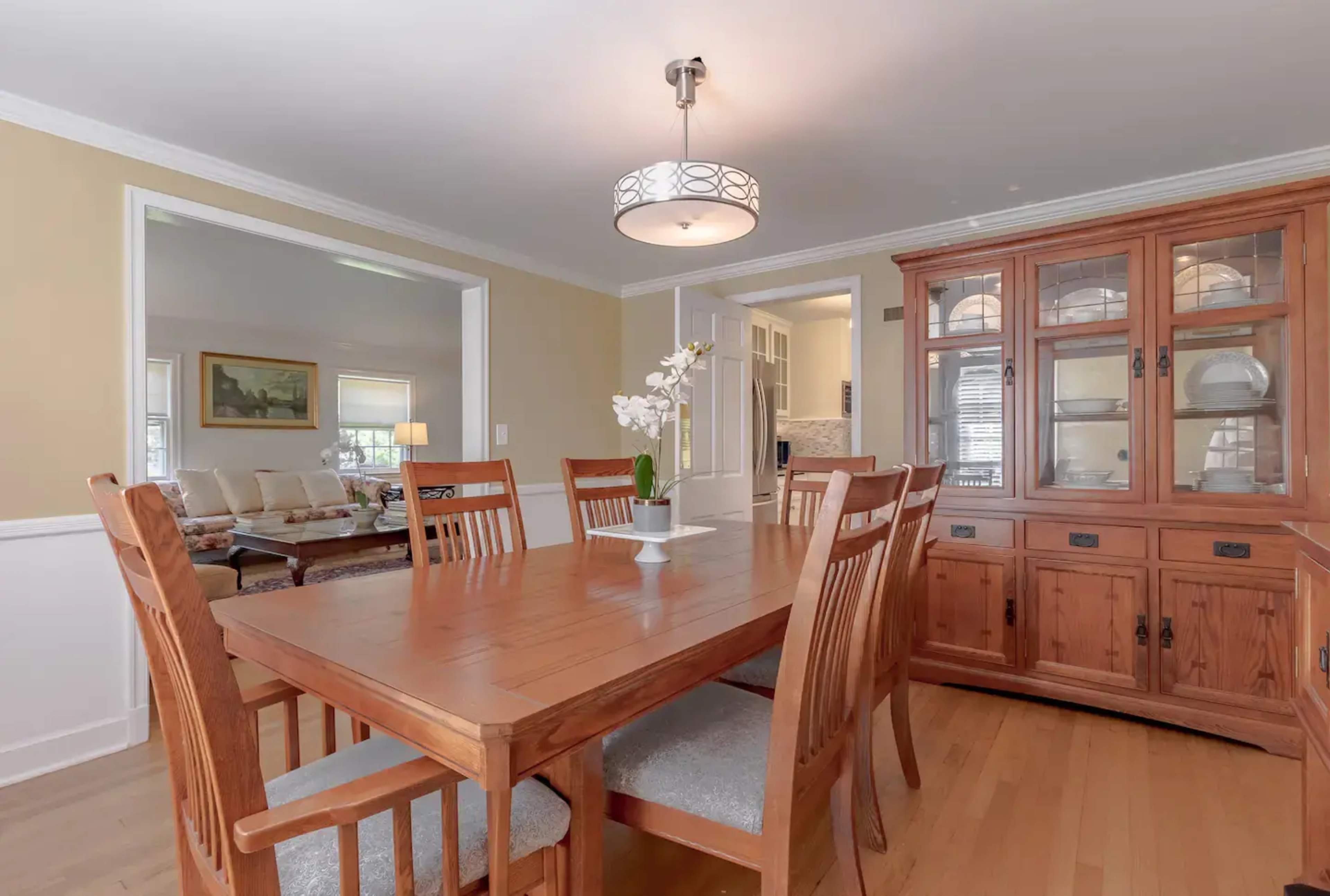 A wooden dining table with chairs is set in a well-lit room featuring a china cabinet and a pendant light.