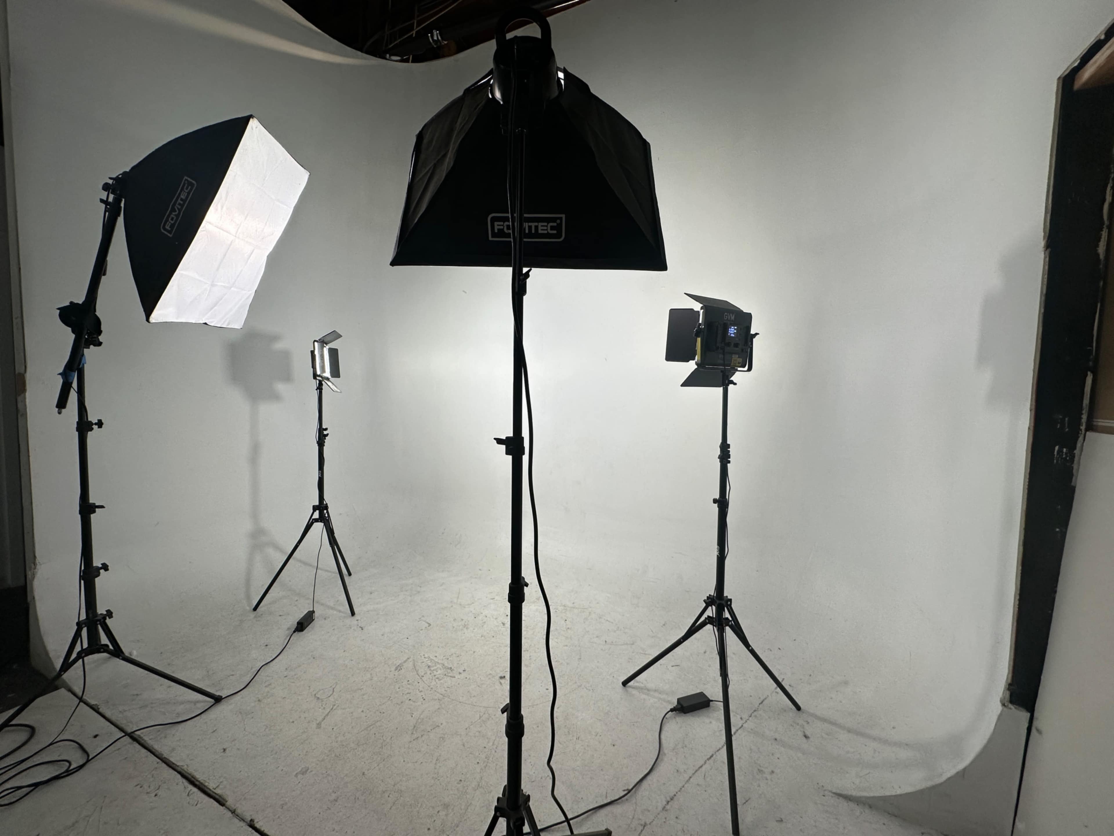 The image shows a photography studio setup with three lighting fixtures on stands facing a blank, white backdrop.