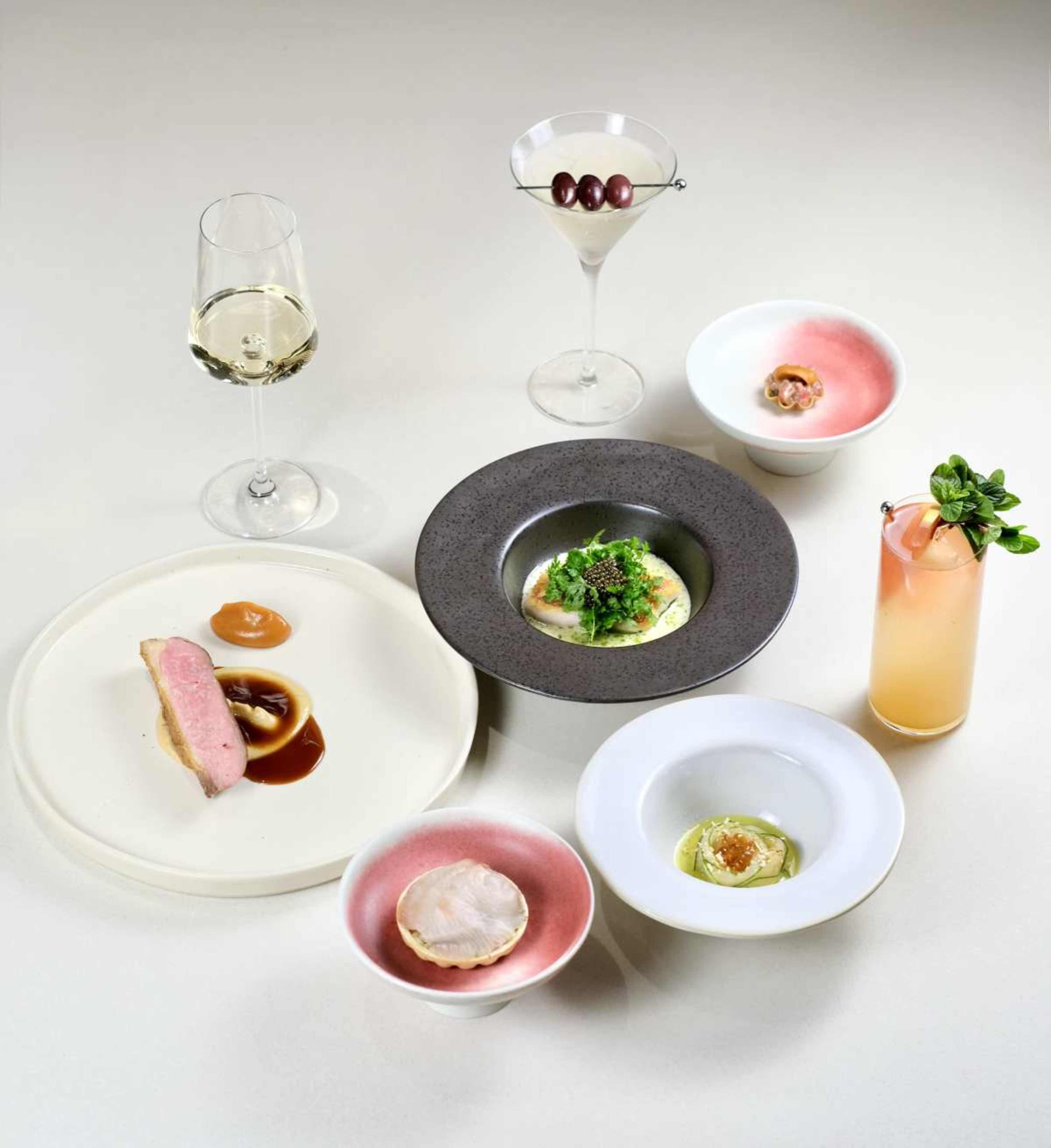 The image displays an elegantly arranged gourmet meal featuring various plated dishes, a cocktail, and a glass of white wine on a neutral background.