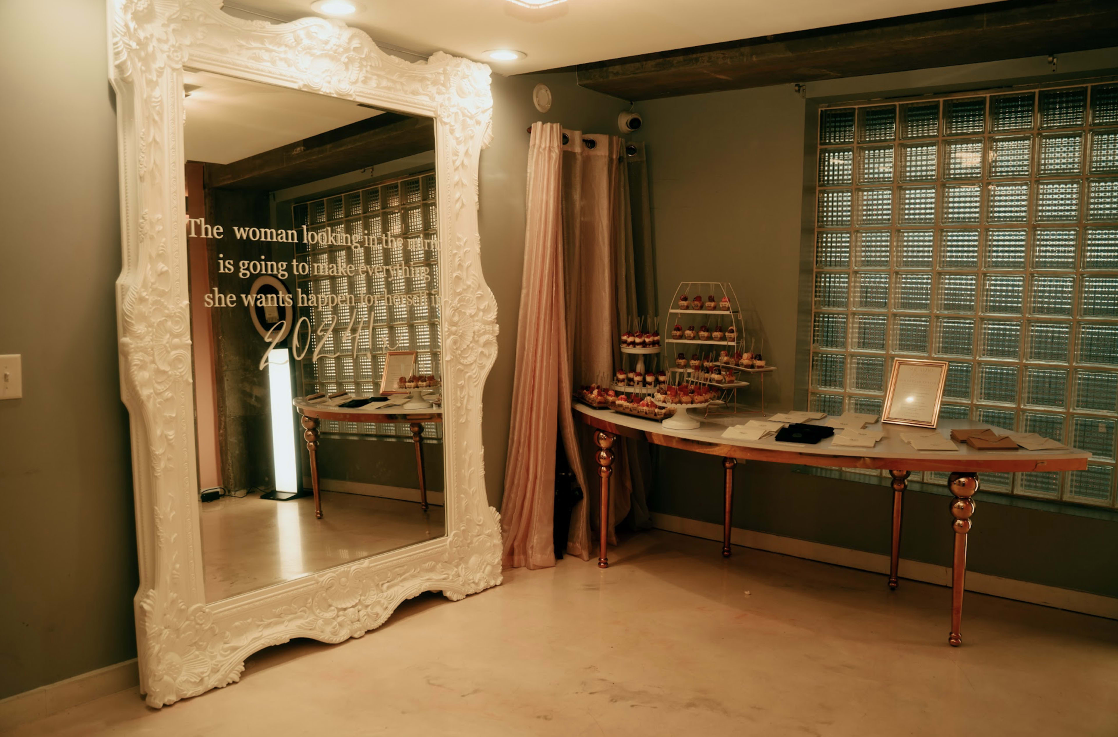 The image shows a decorative mirror on the wall beside a table displaying a selection of desserts in a softly lit room.