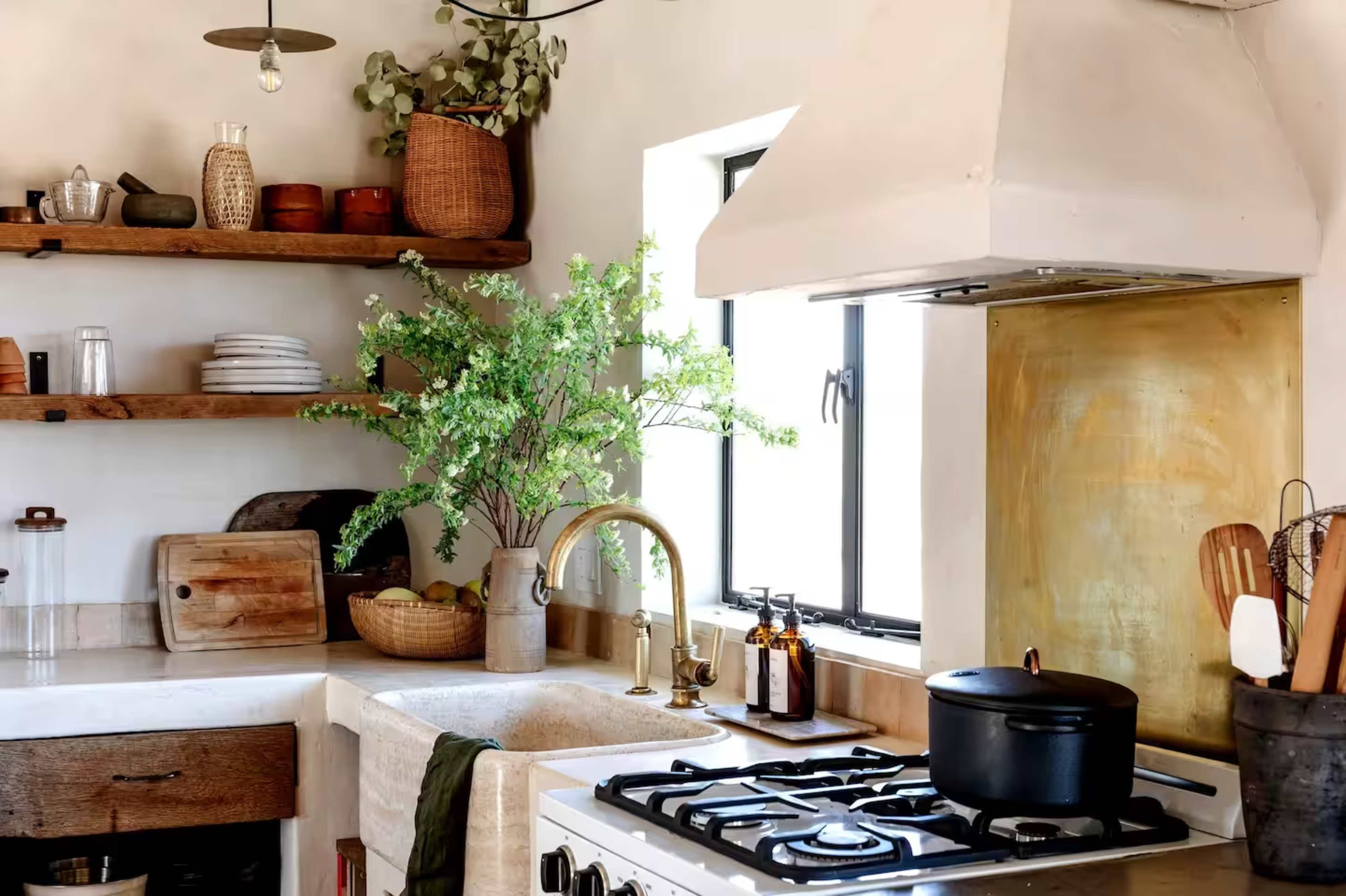 The image shows a modern kitchen featuring open wooden shelves, a farmhouse sink, and a black pot on a gas stove, with potted plants and kitchenware arranged decoratively.