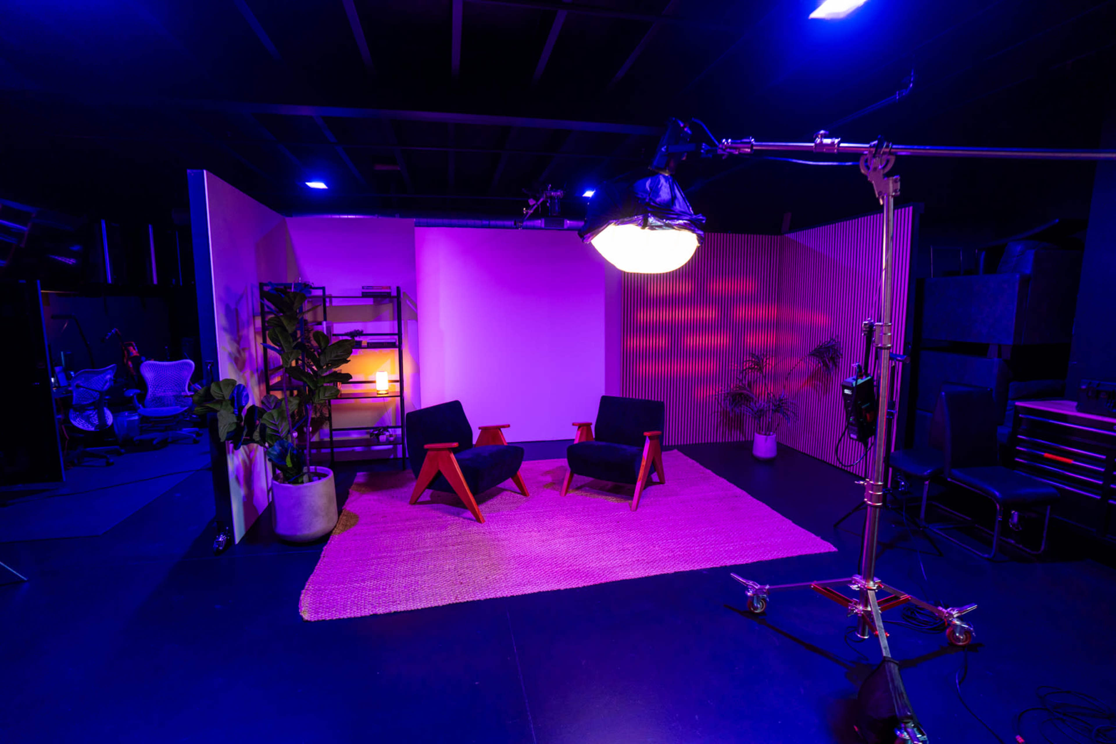 The image shows a stylized studio space with two modern armchairs, a plant, and lighting equipment, all set against vibrant purple walls.