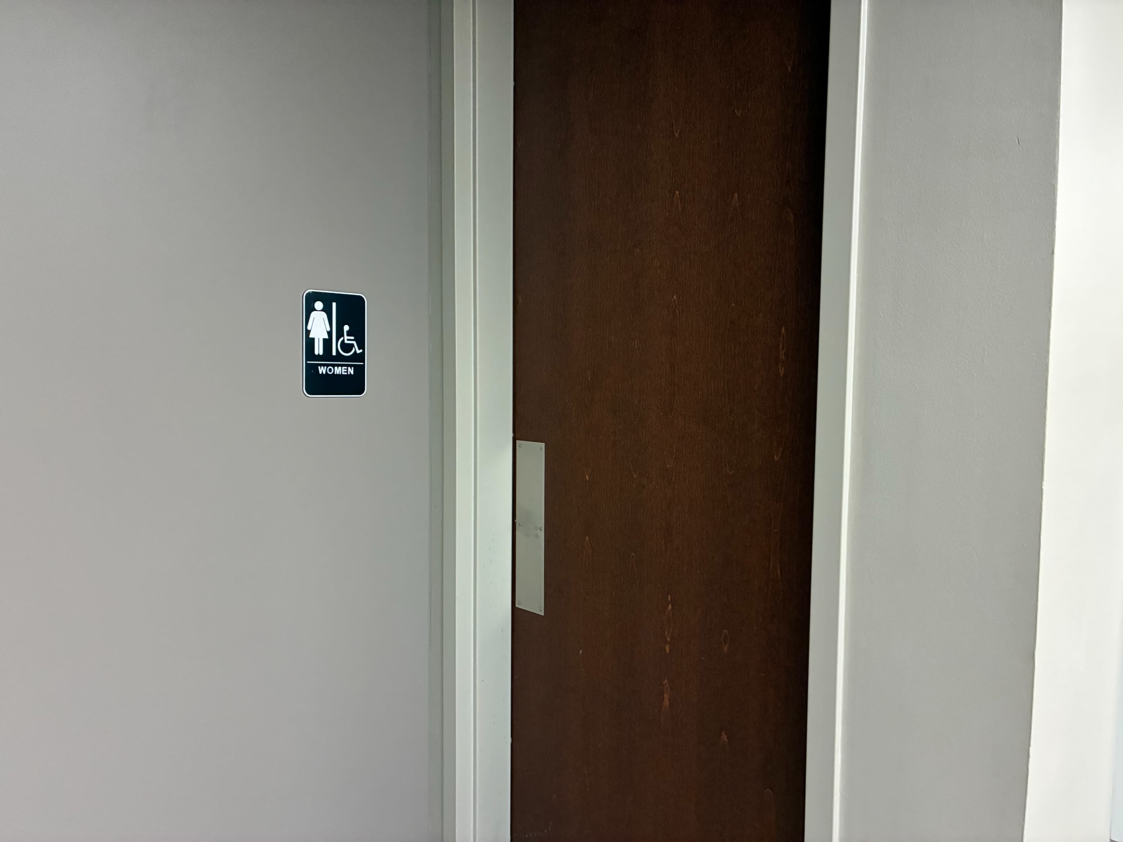 The image shows a women's restroom with a sign indicating accessibility next to a closed wooden door.