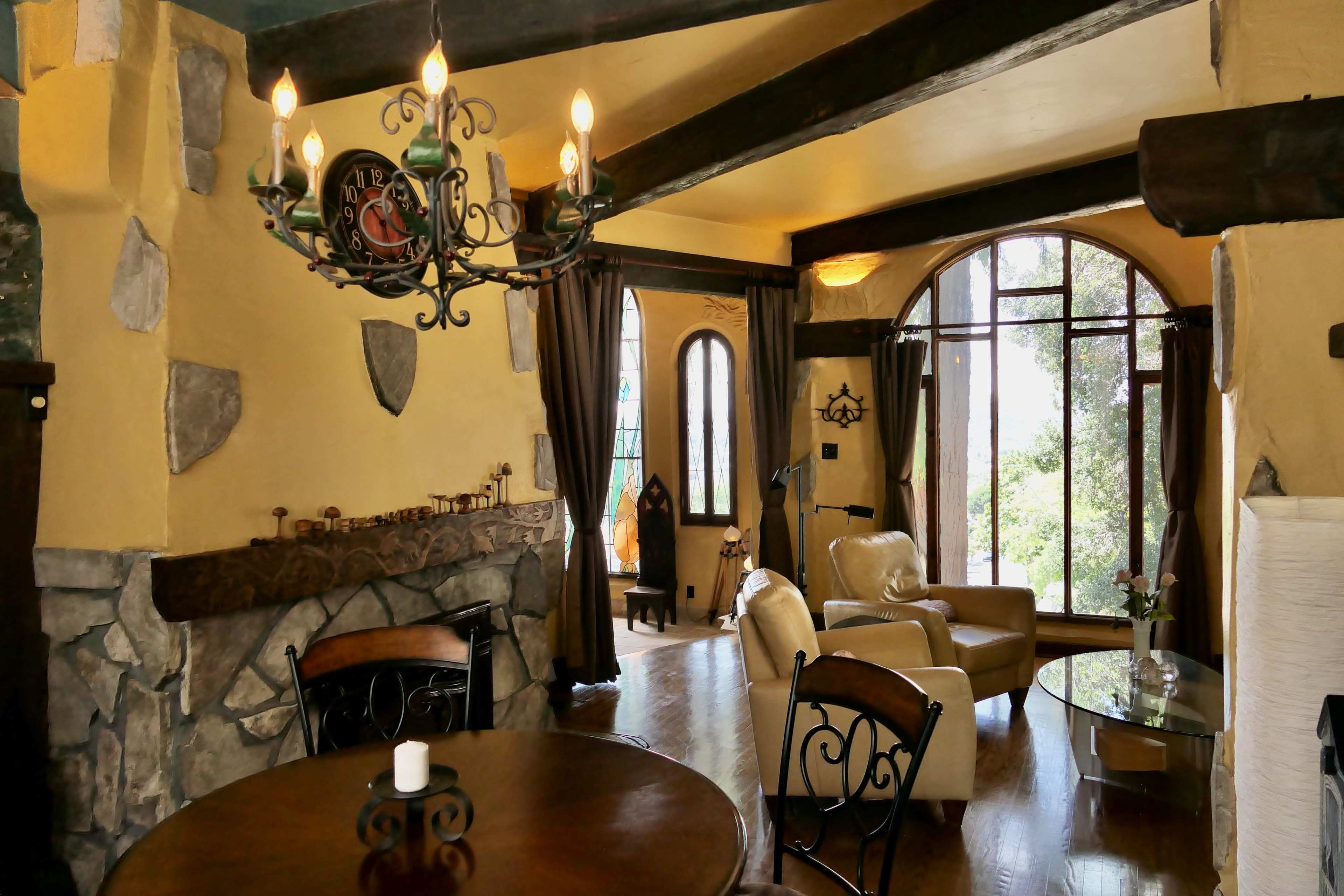 A warmly lit interior living space with a chandelier, a stone fireplace, and large windows overlooking greenery.