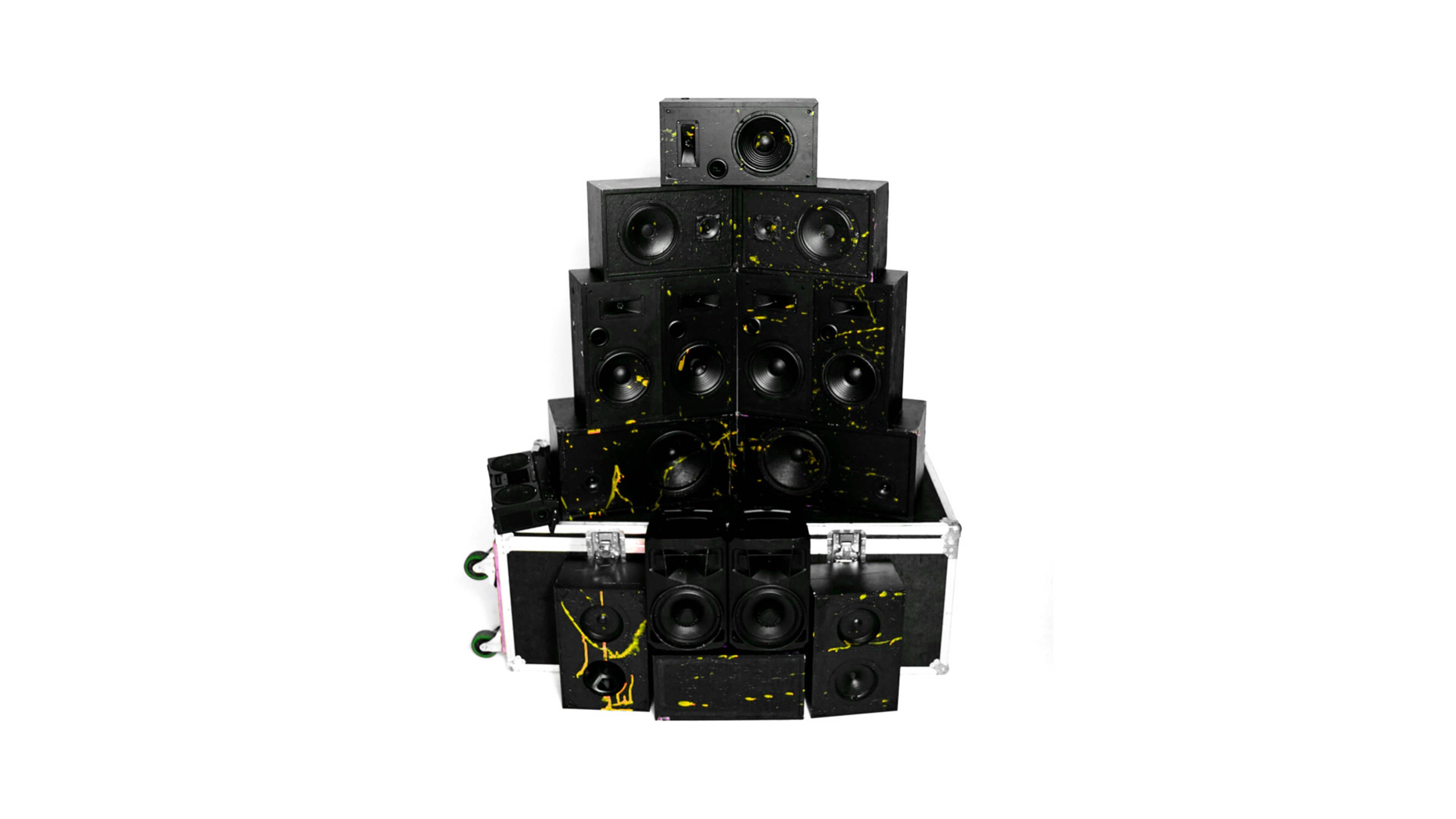 A large arrangement of black audio speakers stacked in a pyramid formation with yellow splatter paint.