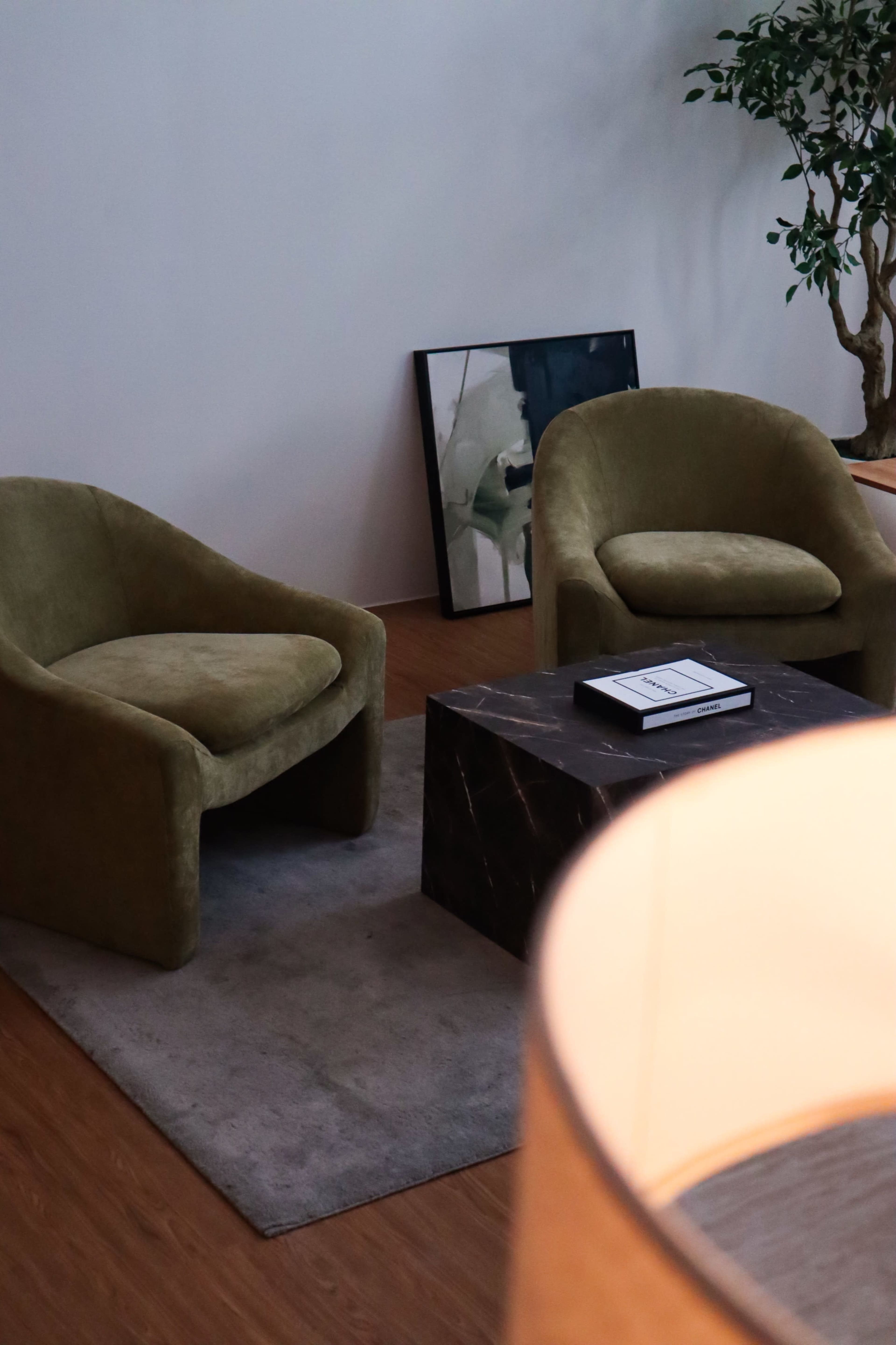The image shows a cozy living room setup with two green armchairs, a black marble coffee table, a decorative painting on the floor, and a floor lamp in the foreground.