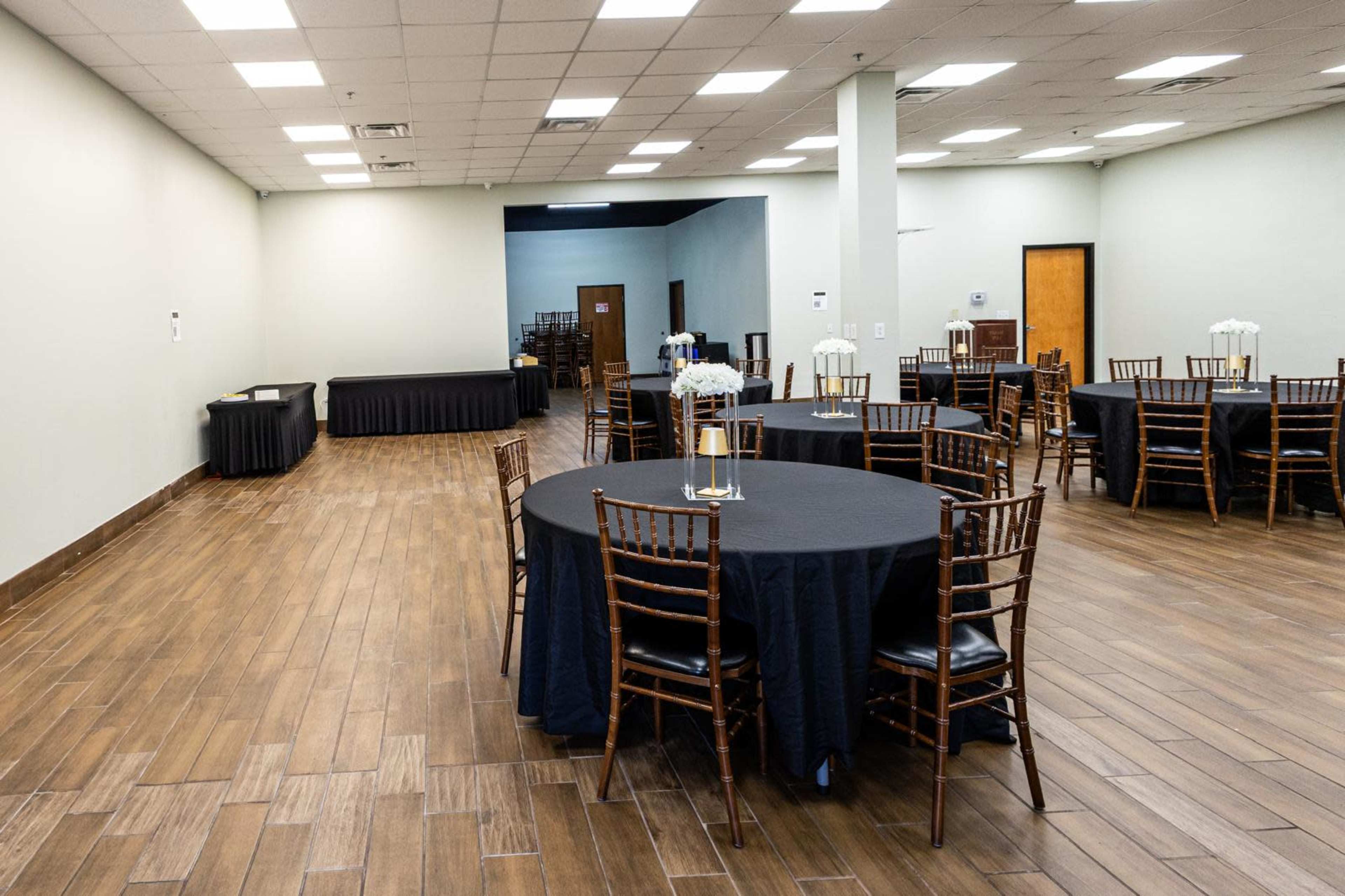 The image shows a spacious event room with several round tables covered in black tablecloths and wooden chairs arranged neatly on a wooden floor.