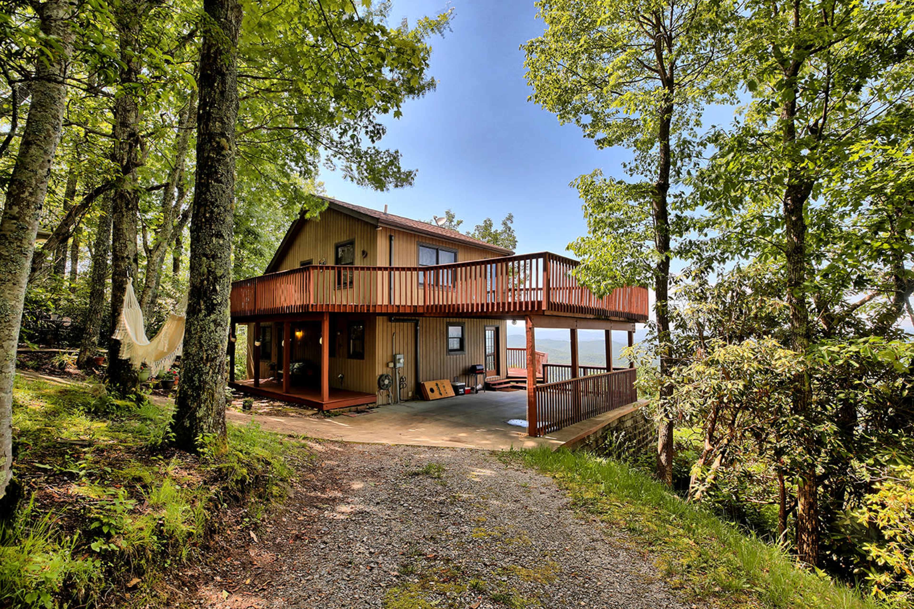 A two-story house with a wooden deck is set among trees, overlooking a body of water in a tranquil outdoor setting.