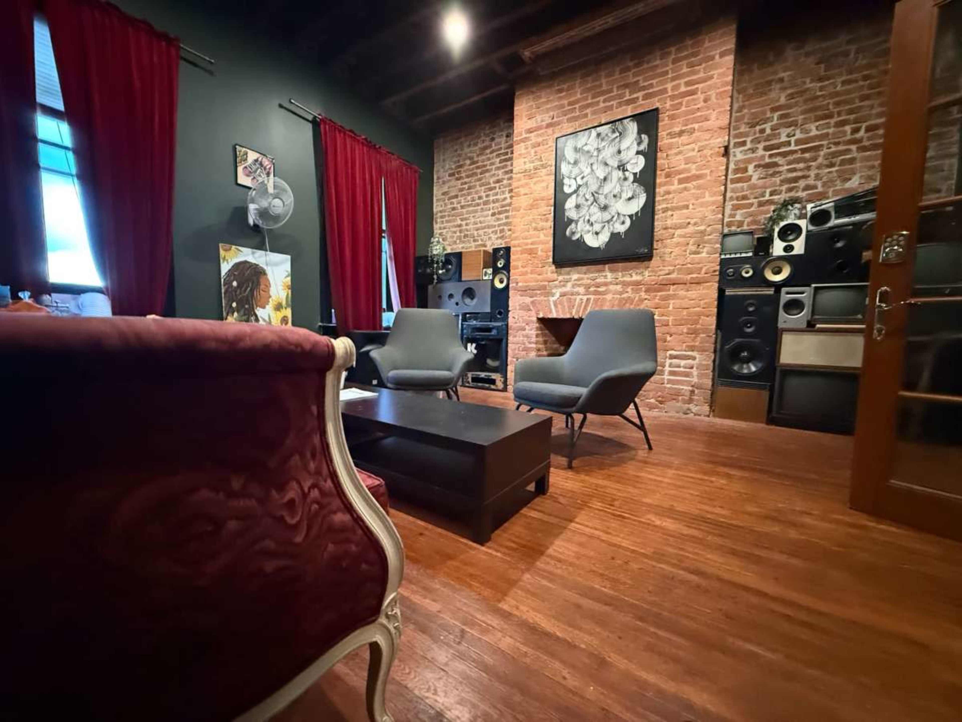 The image shows a cozy interior space with exposed brick walls, red curtains, two modern chairs, a low coffee table, and a collection of vintage speakers on display.