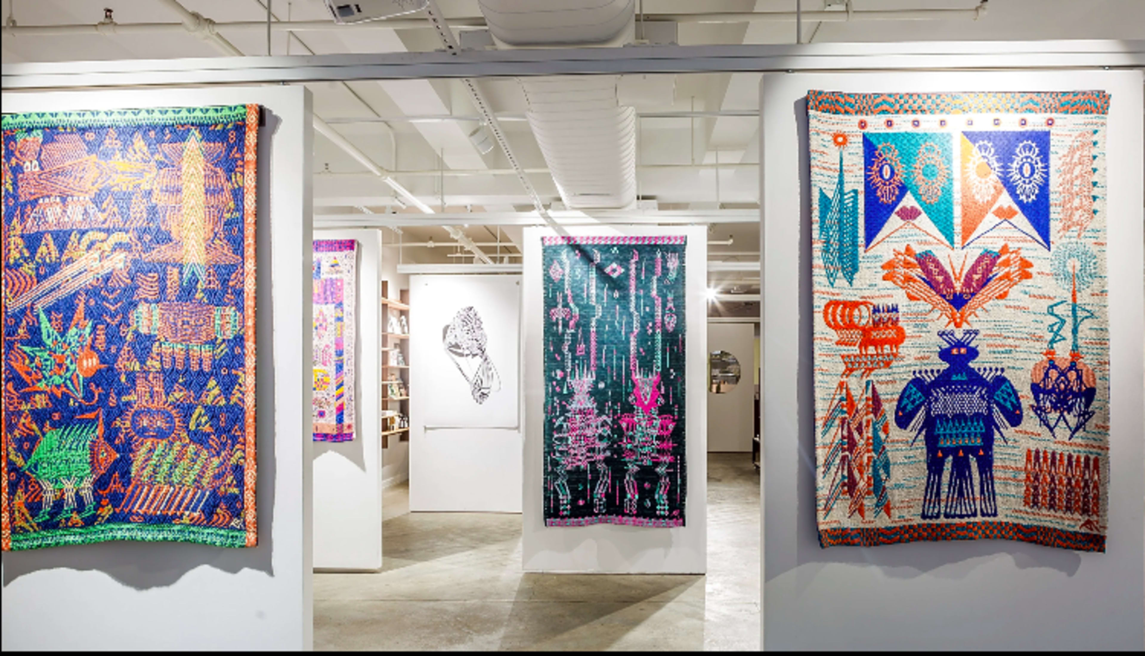 An art gallery featuring colorful woven tapestries displayed on white walls, with additional artwork visible in the background.