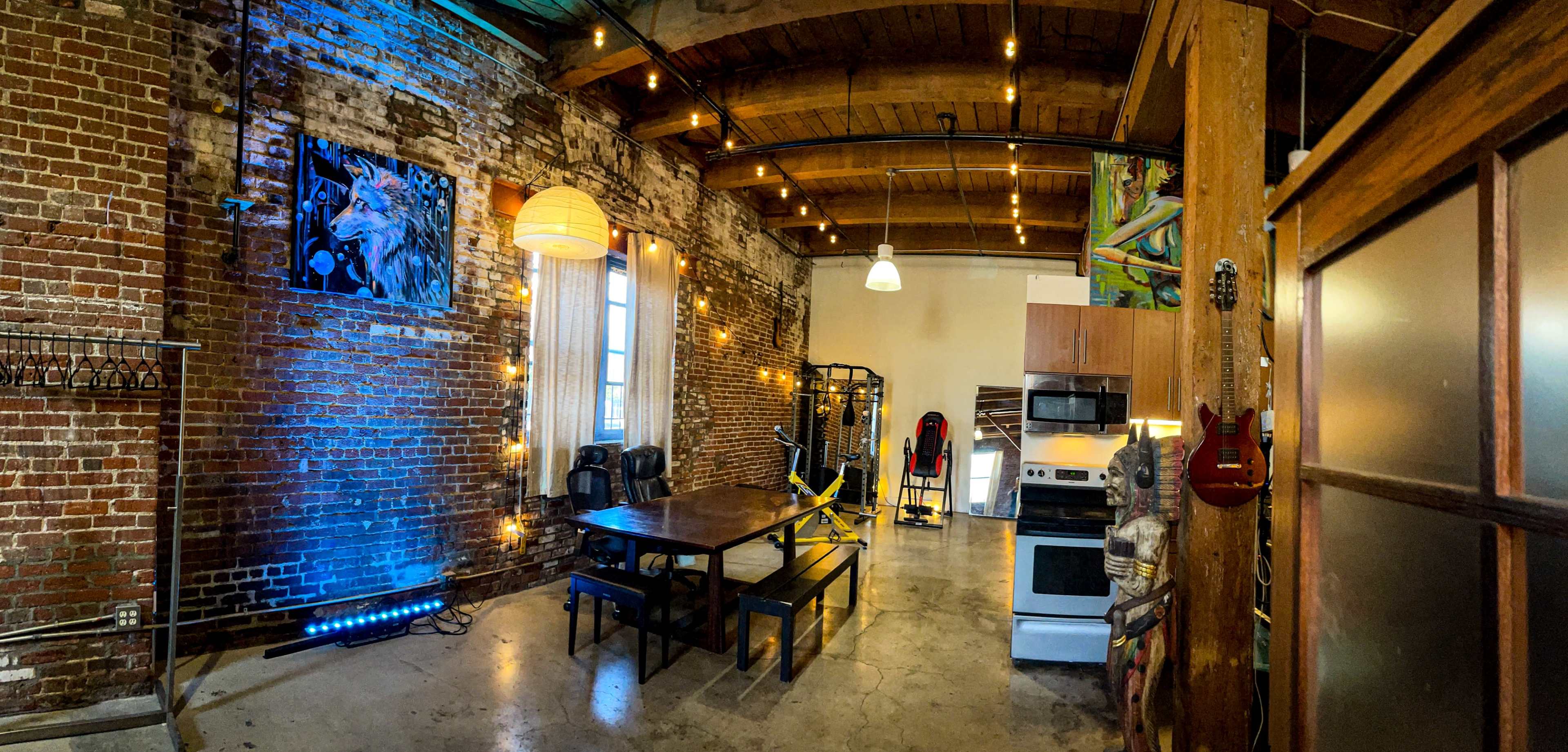An industrial-style loft with exposed brick walls, wooden beams, a large dining table, and kitchen appliances.