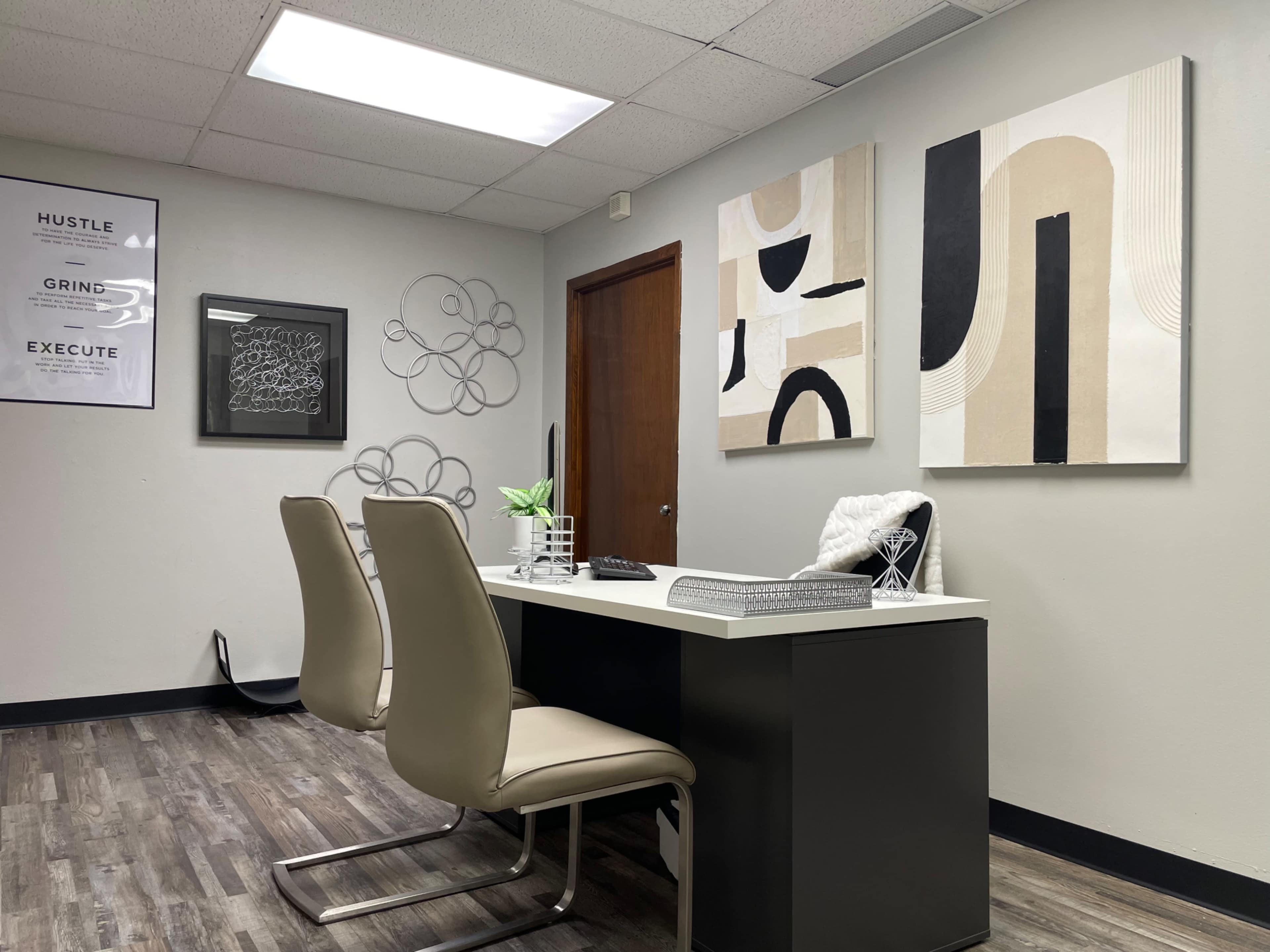 The image shows a modern office space featuring a desk with two chairs, decorative wall art, and a closed wooden door in the background.