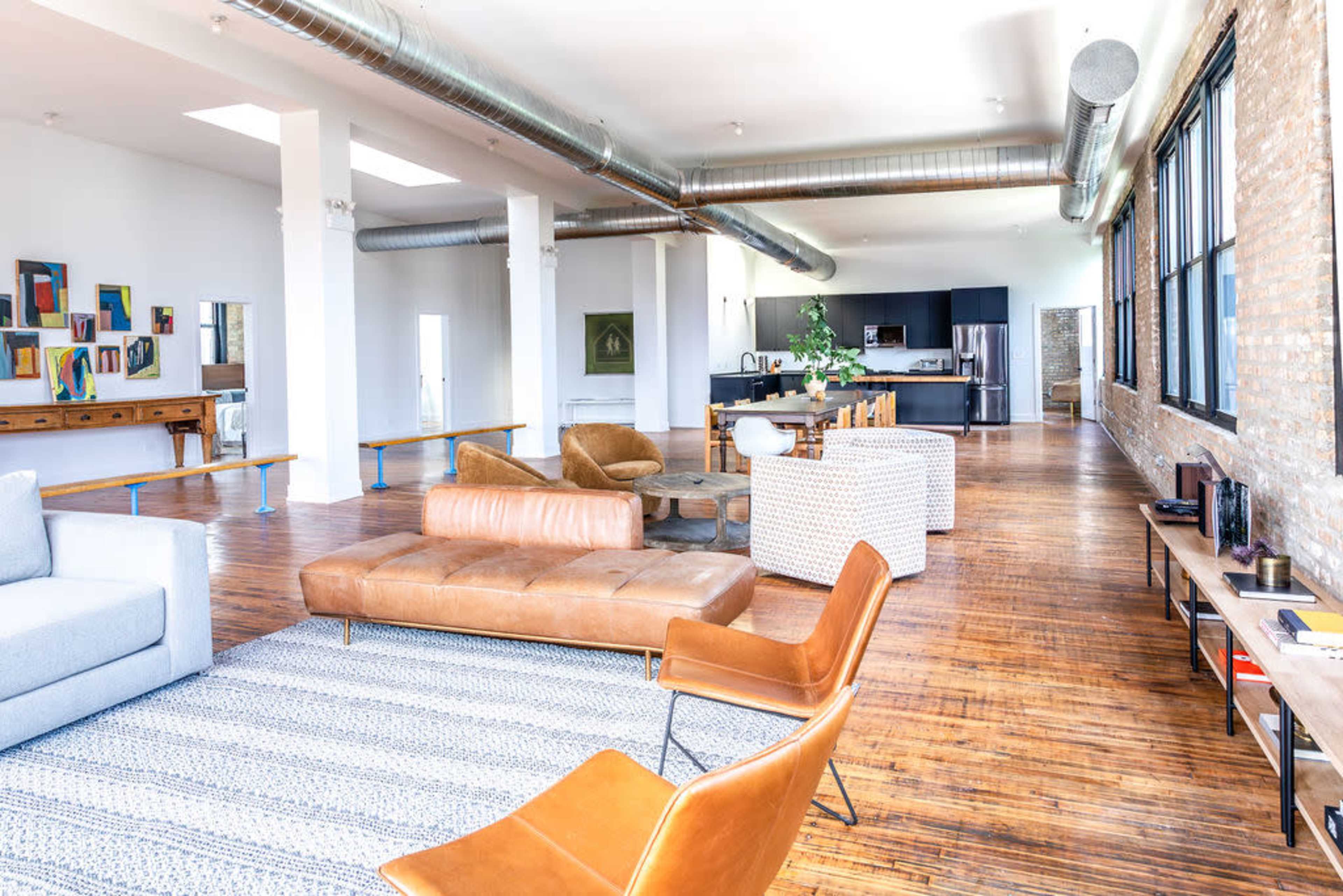 A spacious open-plan living area featuring a mix of modern and vintage furniture, with a kitchen visible in the background and large windows letting in natural light.