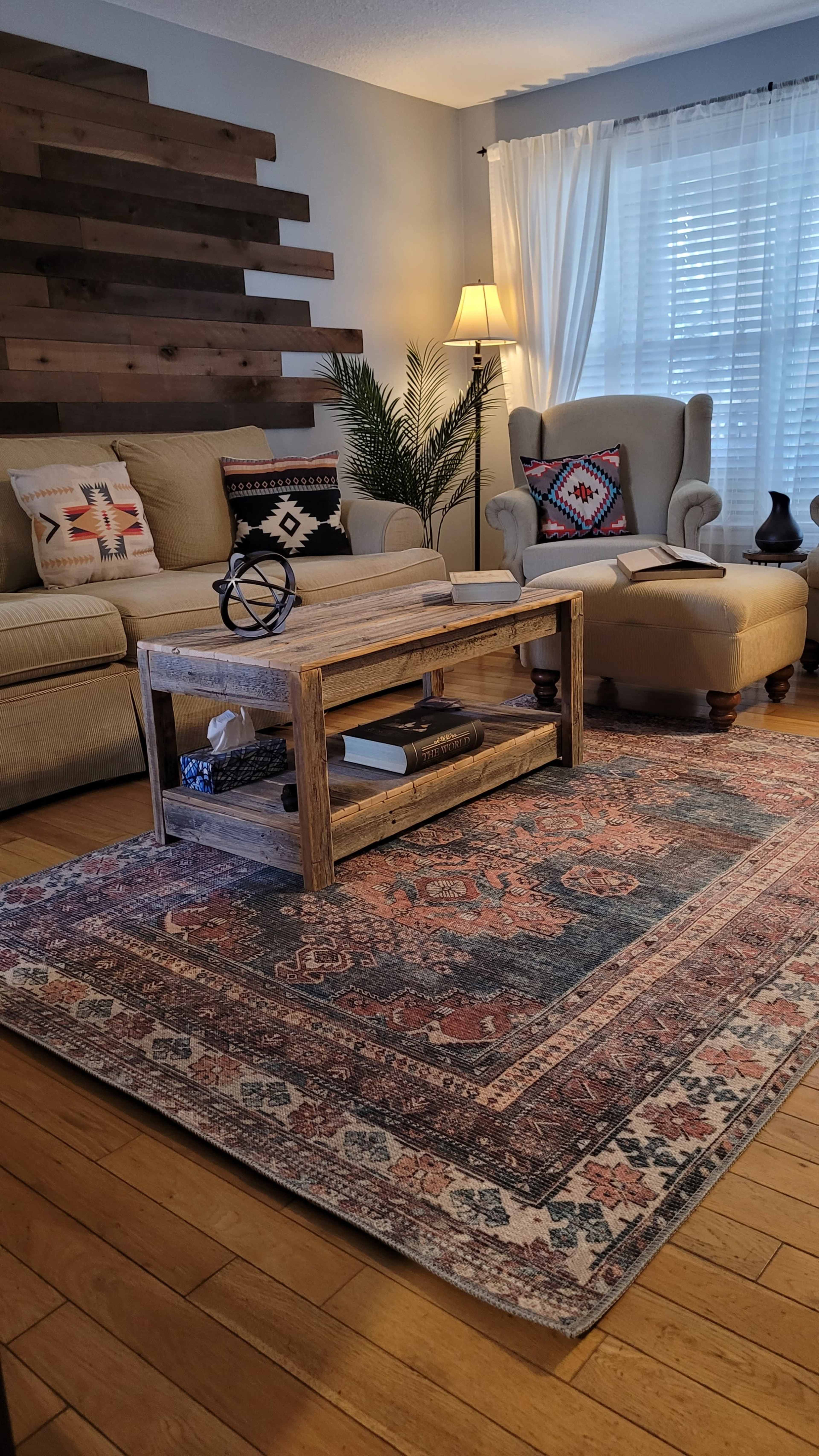 A living room features a beige couch and an armchair, with a wooden coffee table on a patterned area rug, surrounded by decorative pillows and a floor lamp.