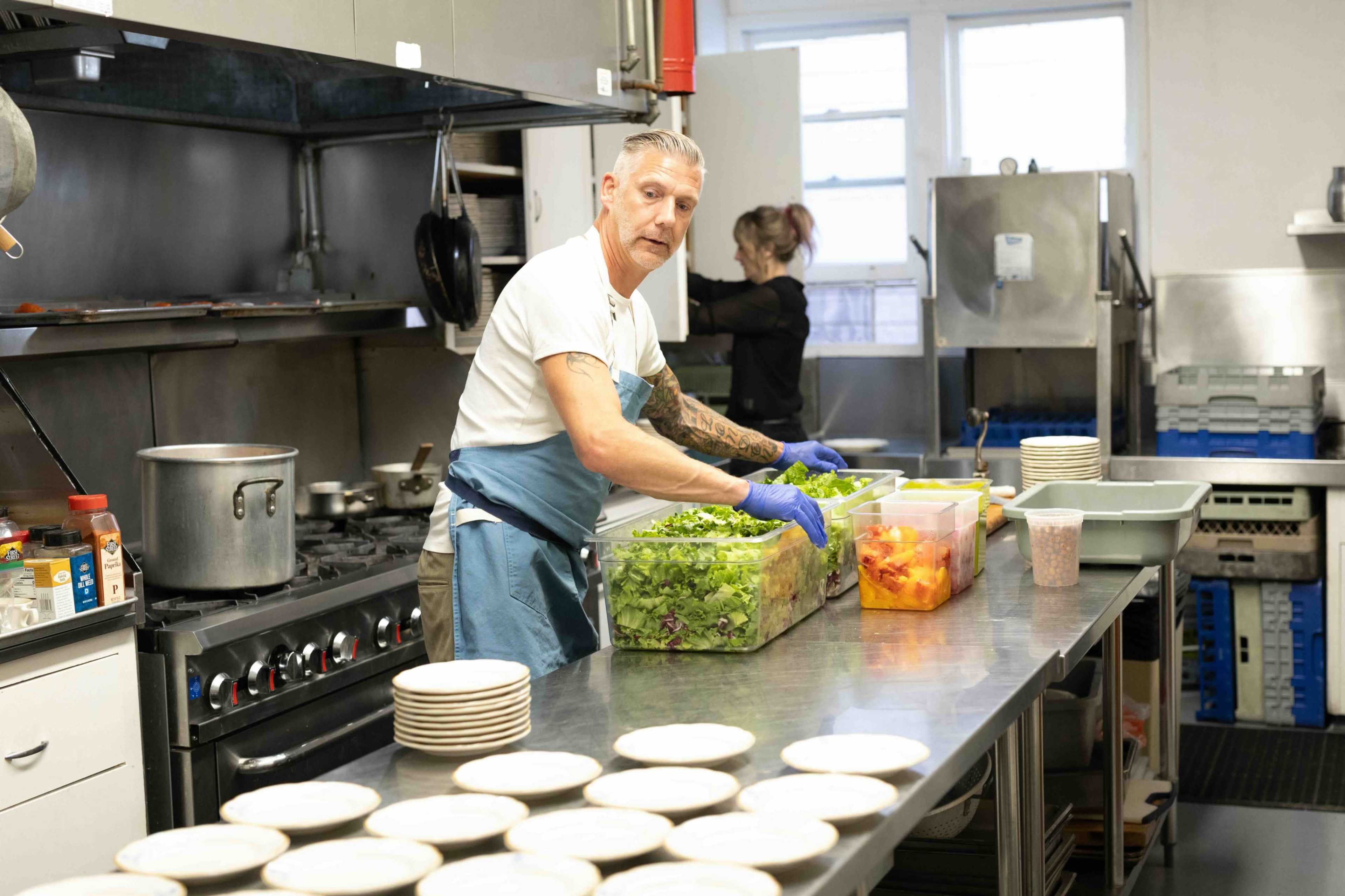 A man prepares fresh salad greens in a kitchen while a woman works in the background.