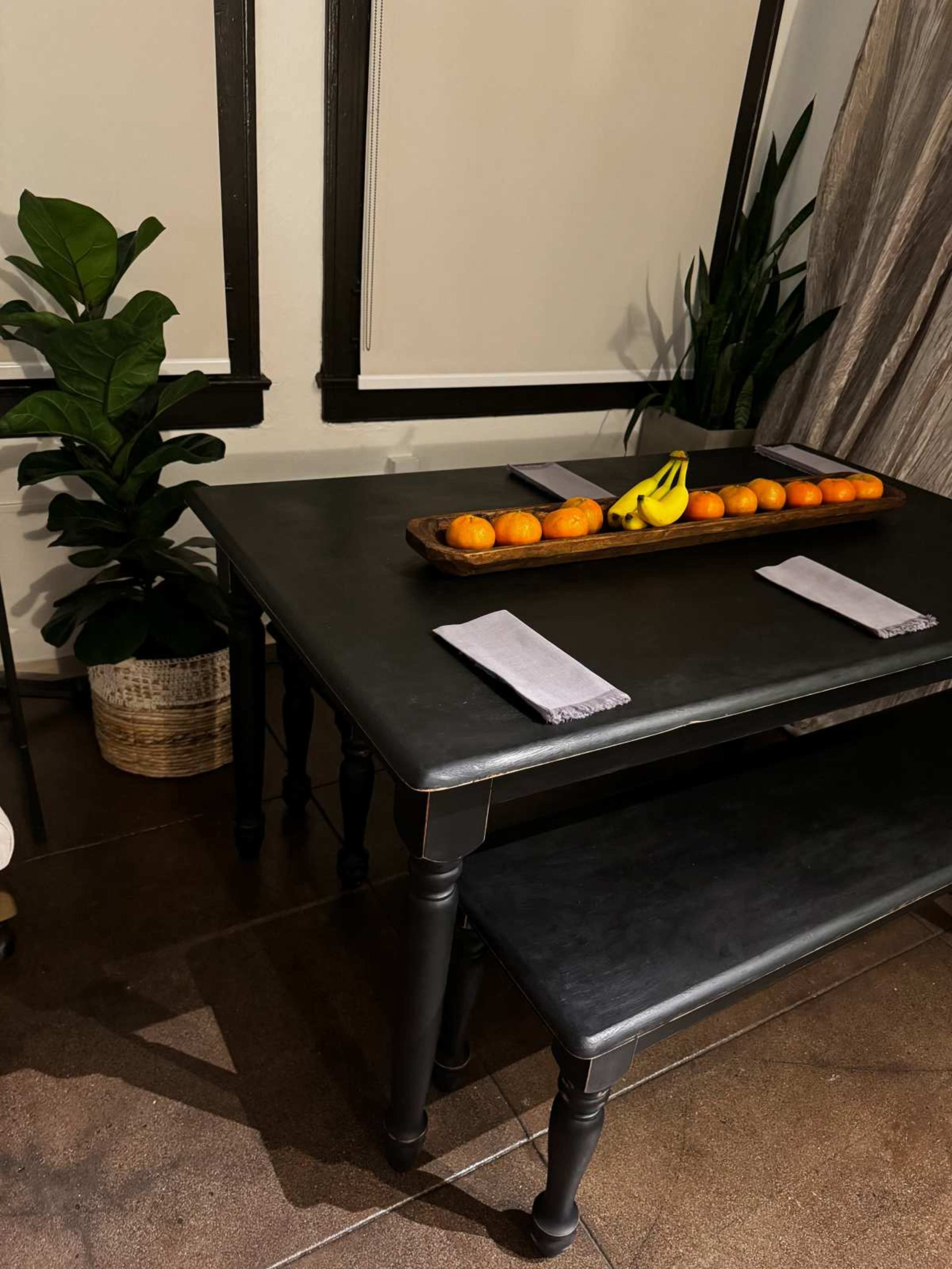 A dark table with a wooden tray of oranges and bananas sits under window coverings, accompanied by a potted plant in the corner.